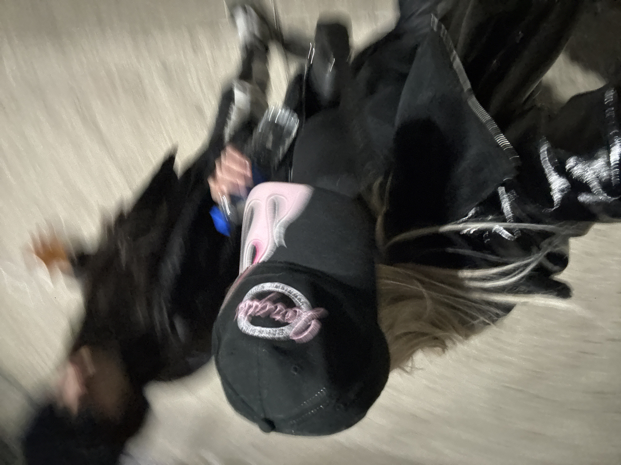 A blurry photograph from a high-angle perspective features a person wearing a black cap with pink embroidery and pink flame designs. The individual is dressed in dark clothing, including a black jacket. A plastic water bottle is visible near the person's hand. Part of their blonde hair is visible beneath the cap. The background consists of a light-colored, textured surface, which may be concrete or a similar material. The location is unknown, but could theoretically be in Buenos Aires, Argentina.