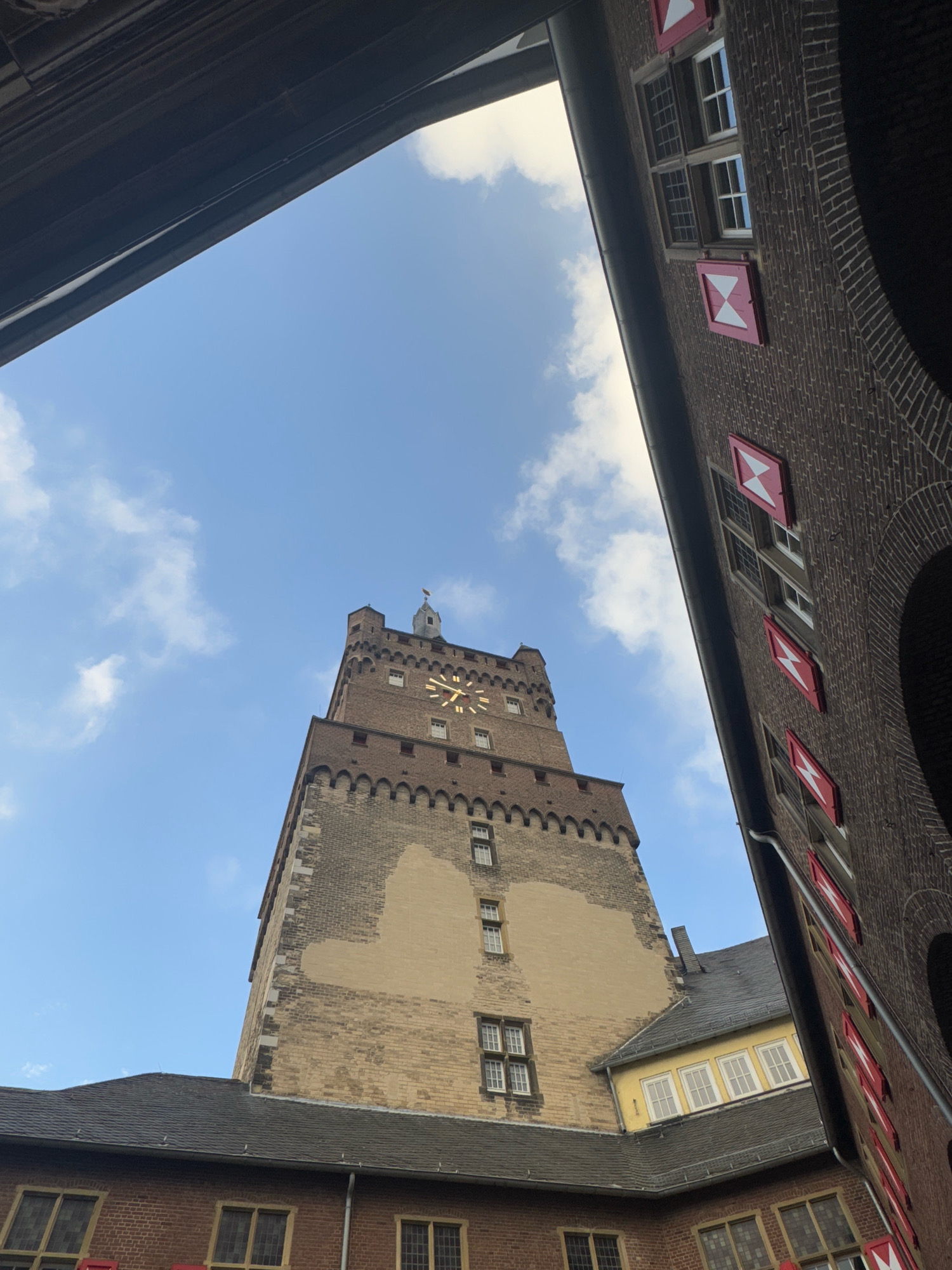 The image is a low-angle shot of the Cleves Castle Tower in Cleves, Germany. The tower, constructed from brick and stone, features a large clock face. Red and white shutters adorn the windows of the adjacent brick buildings, which are partially visible on the right and in the foreground. The sky is blue with scattered white clouds. The scene appears to be an outdoor courtyard or open space.
