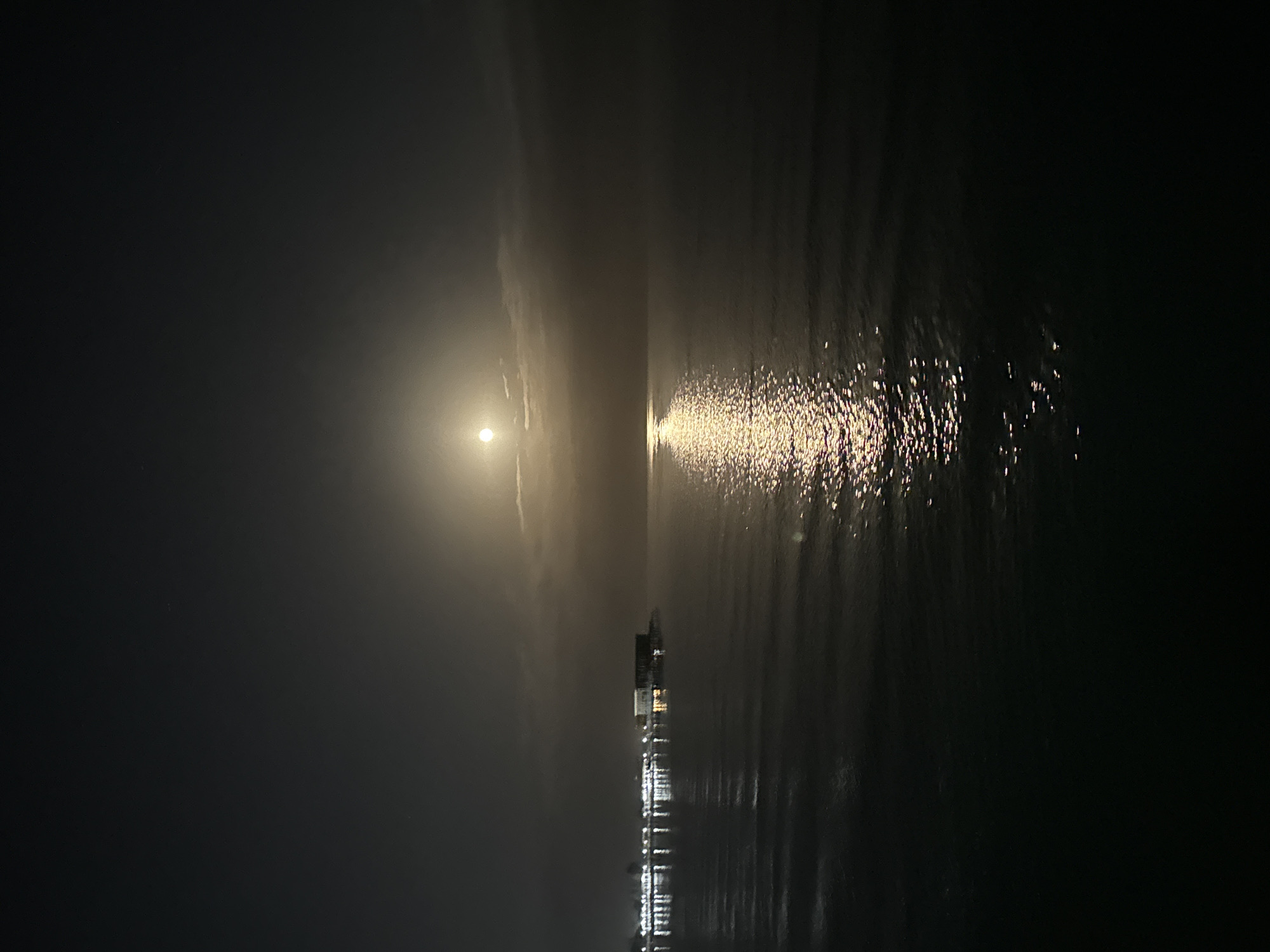 The image depicts a nighttime scene of a body of water, likely Issyk-Kul Lake, with a full moon visible in the sky. The moon's light casts a shimmering reflection on the calm water's surface. In the distance, along the shoreline, there is a line of lights that appear to be from buildings or a pier, suggesting a developed area. The overall atmosphere is serene and tranquil, illuminated by the natural light of the moon and the artificial lights on the shore.