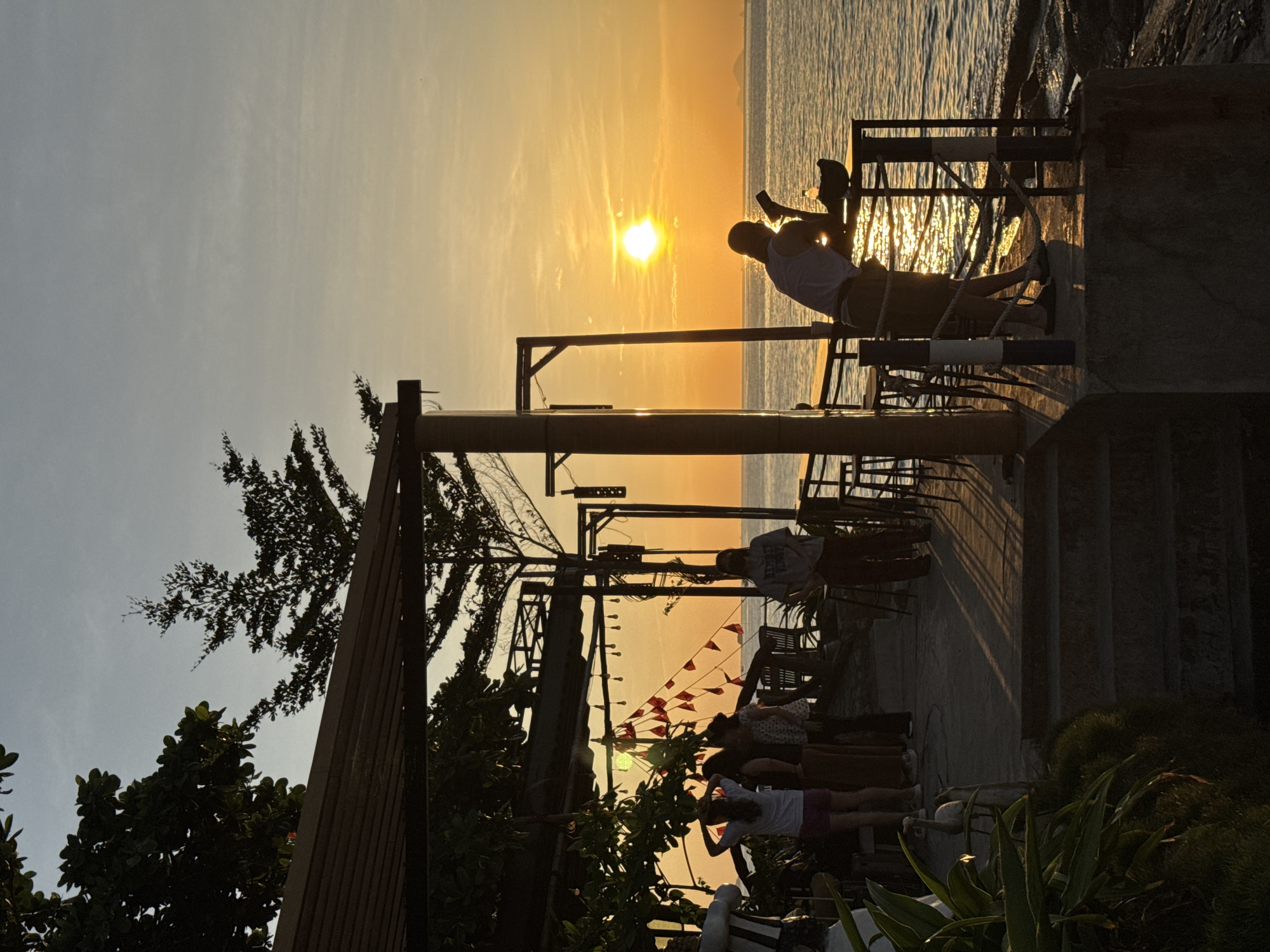 The image depicts a sunset scene at a waterfront establishment in Nha Trang, Vietnam. A person is silhouetted against the bright orange sky, leaning on a railing with a drink in hand. Several other individuals are visible in the background, some standing and others seated at tables. Red flags are strung across a canopy above them. The establishment appears to be situated on an elevated platform overlooking the sea, with rocky shorelines visible in the foreground. The golden light of the setting sun casts long shadows across the patio.