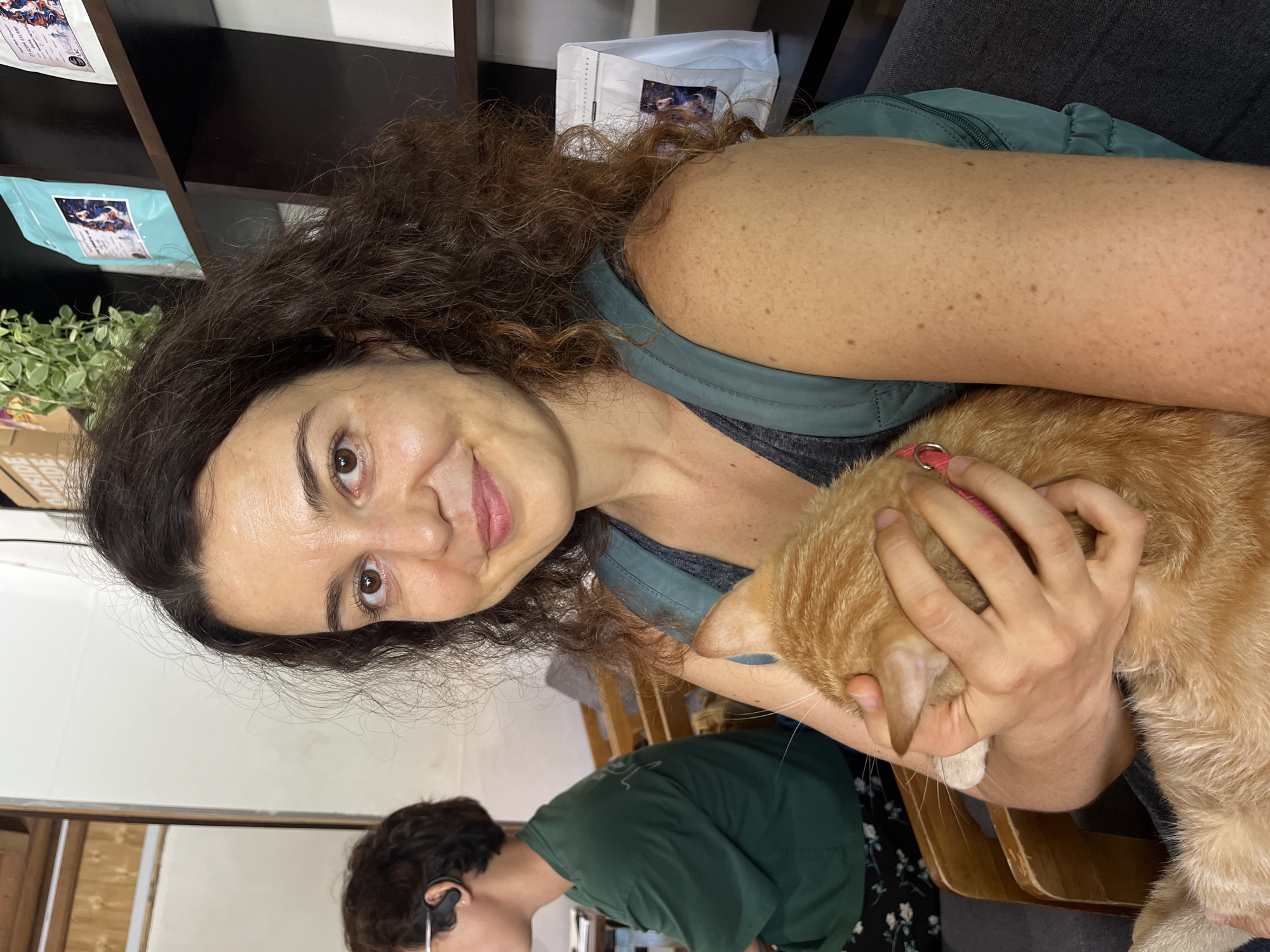 A woman with curly brown hair is holding an orange tabby cat in her arms. She is smiling and looking at the camera. The cat is also looking away from the camera. In the background, another person is seated, wearing a green shirt. The setting appears to be indoors, possibly a cafe or a shop, with shelves of products visible on the right side of the image. The overall lighting is bright and natural.