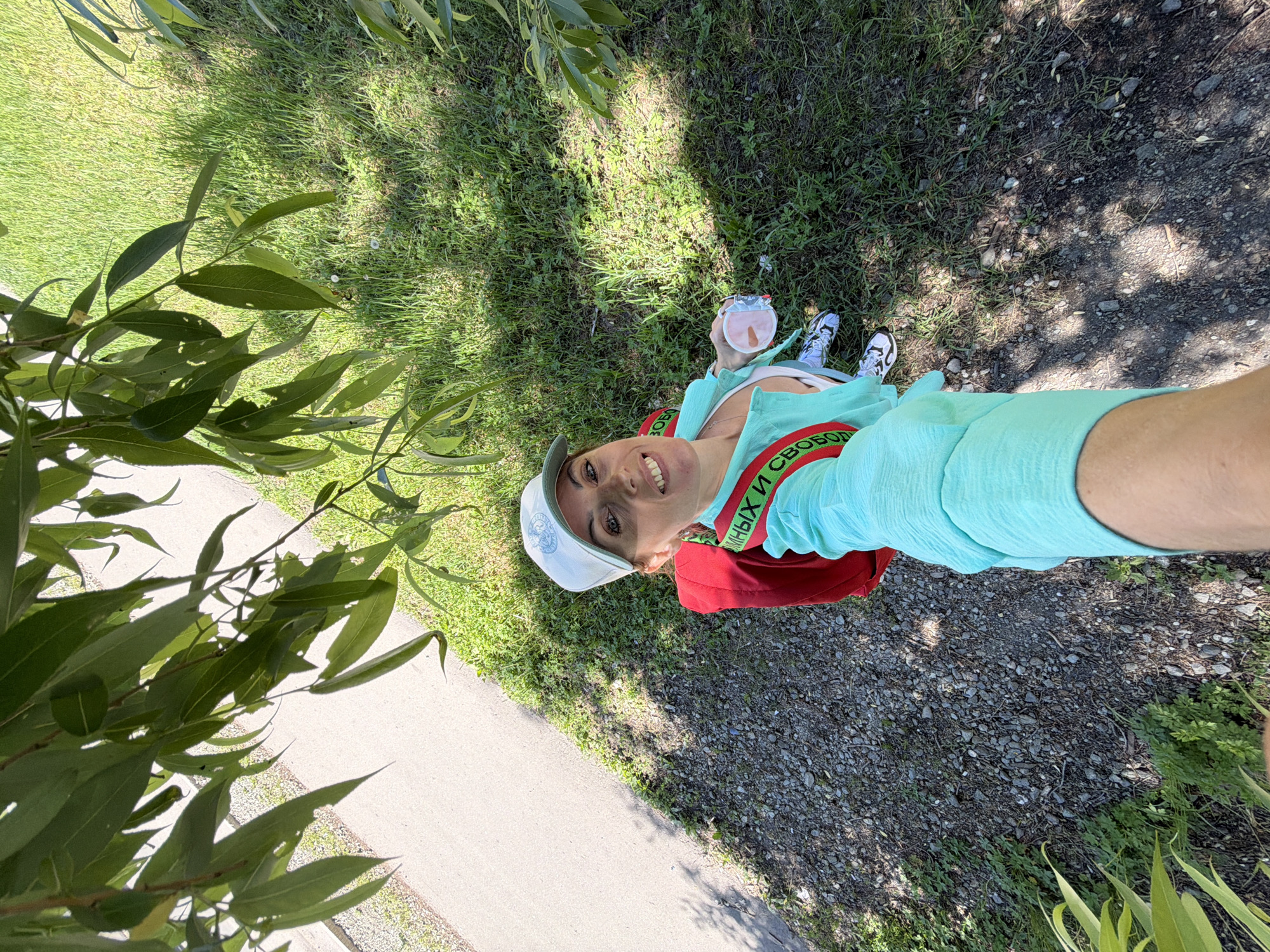 A woman is taking a selfie from a high angle under a tree in Birobidzhan, Russia. She is wearing a light blue baseball cap, a turquoise shirt, and a red backpack. Her left hand is holding a clear cup with a pink lid. She is smiling at the camera. The ground around her is a mix of gravel and grass, with a paved path visible to the left. Sunlight is filtering through the tree leaves, creating shadows on the ground.
