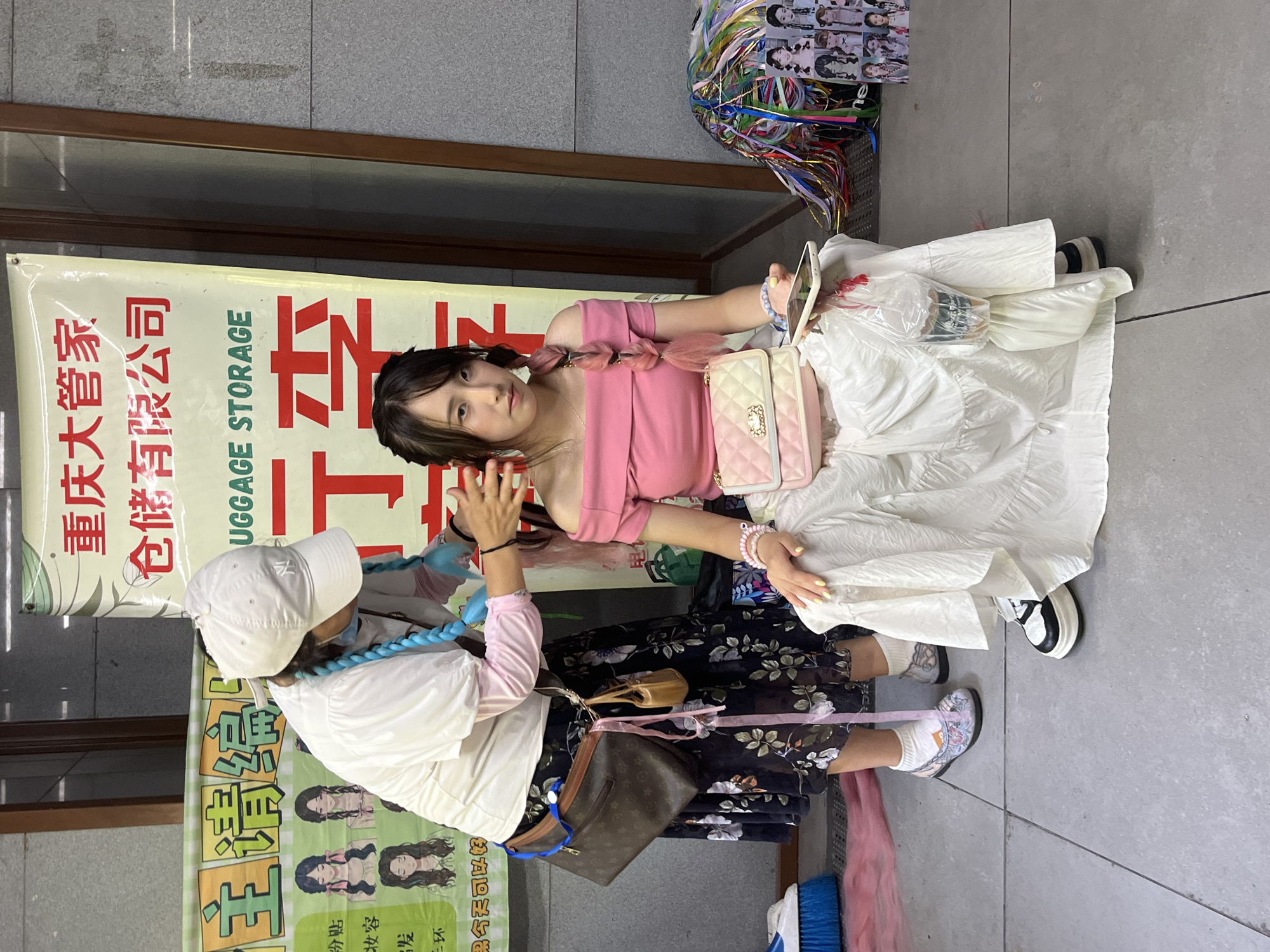 A young East Asian woman with pink-tinted braided hair sits on a grey tiled surface, looking at the camera with a neutral expression. She is wearing a pink off-the-shoulder top and a long white tiered skirt. She holds a smartphone and a plastic cup with a straw in her right hand, and a pastel pink quilted handbag rests on her lap. To her left, another person, likely a woman, wearing a white cap, white top, and a dark floral skirt, is braiding the seated woman's hair. The braider has bright blue braided hair extensions visible. A large banner hangs behind them, with Chinese text at the top reading "重庆大管家, 仓储有限公司" (Chongqing Big Housekeeper, Warehousing Co., Ltd.) and "UGGGAGE STORAGE" in English. To the left of the banner, another green banner displays cartoon drawings of women with various hairstyles. Scattered on the ground are colorful ribbons, a blue broom, and several small printed images of women with braided hairstyles. The scene appears to be outdoors in an urban setting, possibly a street market or sidewalk. The lighting suggests it is daytime.