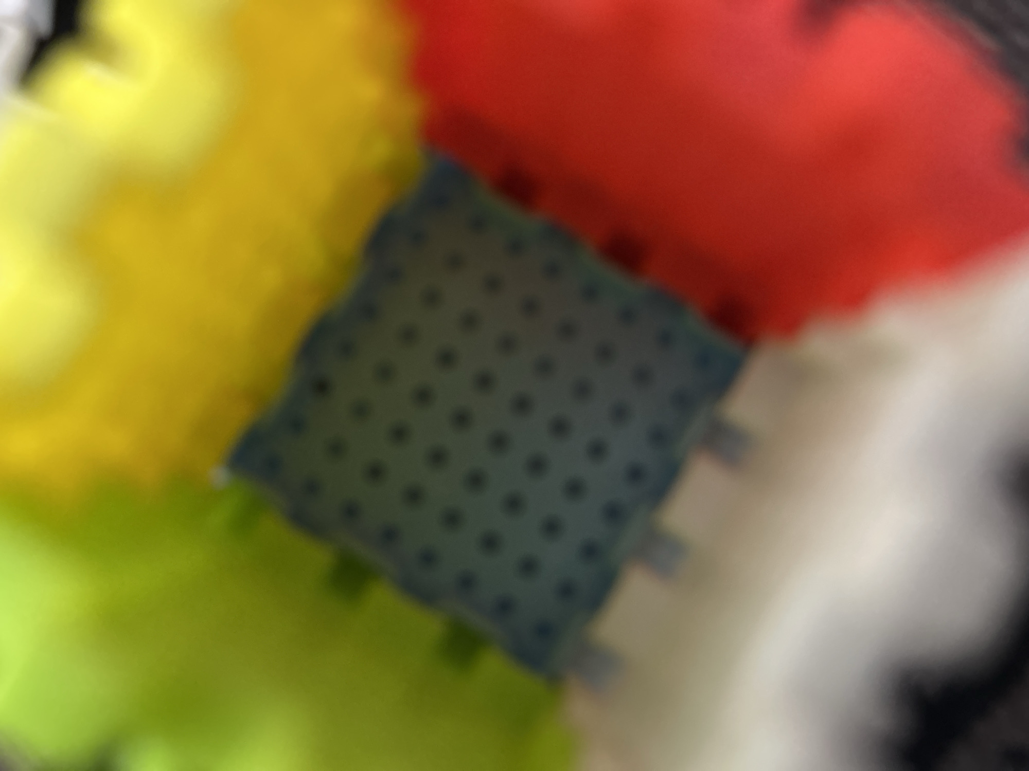 The image shows a close-up, blurred view of what appears to be interlocking building blocks. The blocks are in various bright colors, including yellow, red, green, and white, surrounding a central blue block that has a pattern of small, dark dots. The texture of the blocks is visible, with raised edges and indentations characteristic of interlocking toys. The lighting is diffused, suggesting it might be an indoor setting, and there are no clear indicators of the time of day or weather conditions. The focus is on the arrangement and colors of the blocks, creating an abstract, playful composition. No people are visible, and there is no discernible text within the frame.