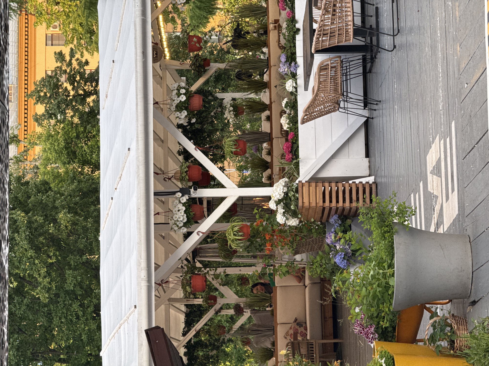 The image shows an outdoor dining area, likely in Moscow, Russia. The space is covered by a white, tent-like structure with wooden beams, decorated with hanging plants in terracotta pots and various flowers. Seating consists of a beige sofa with patterned pillows and several wicker bar stools. The floor is made of gray wooden planks. A large gray planter with flowers and greenery sits at the edge of the dining area. A yellow building can be seen in the background through the trees. People are seated inside.