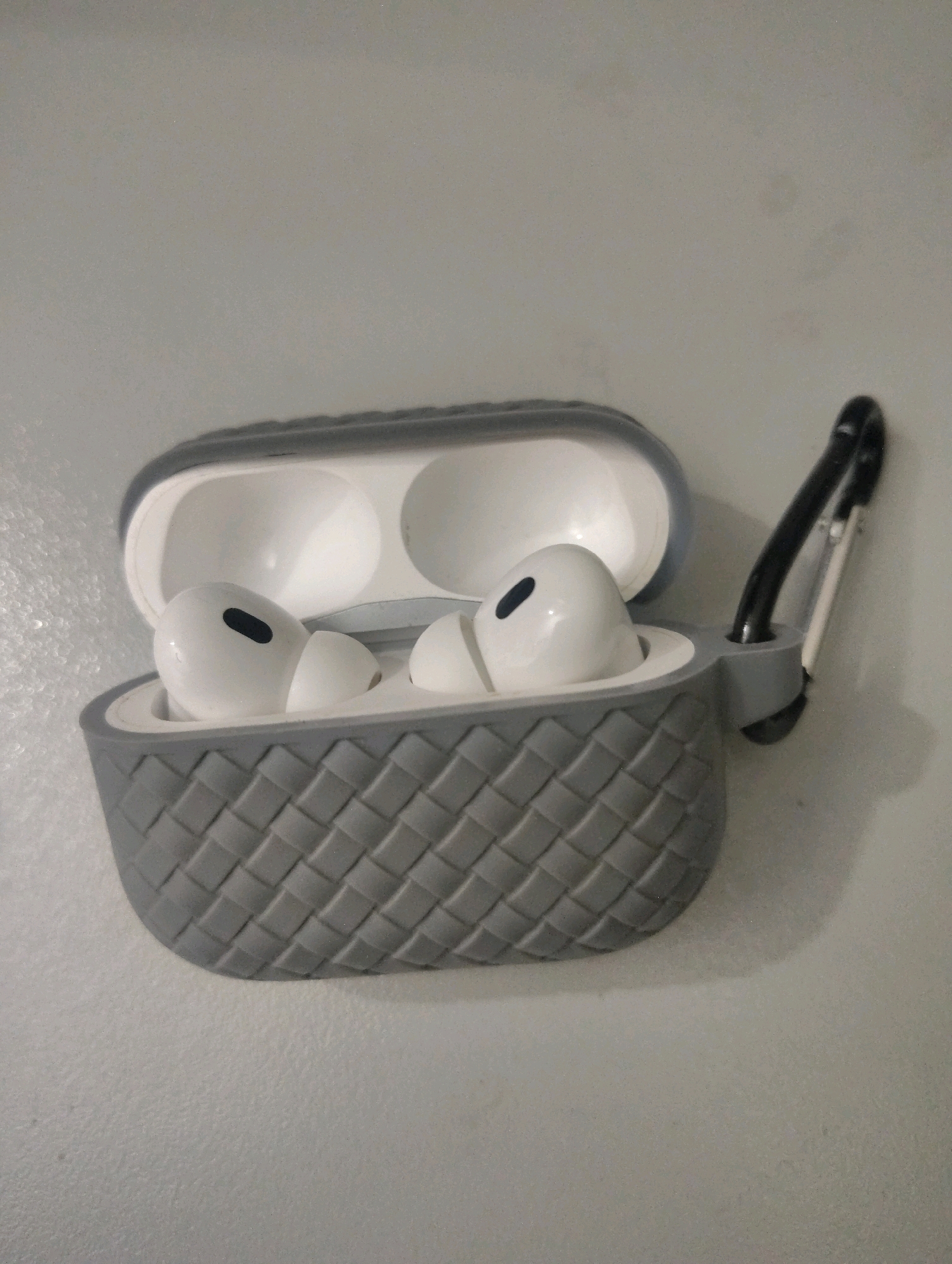 A pair of white wireless earbuds with a black accent are placed inside an open grey charging case with a woven texture. The charging case has a black carabiner attached to its side. The earbuds and case are positioned on a light grey, textured wall. The image does not provide any information or visual cues that would allow for the identification of a specific location in Lahore, Pakistan.