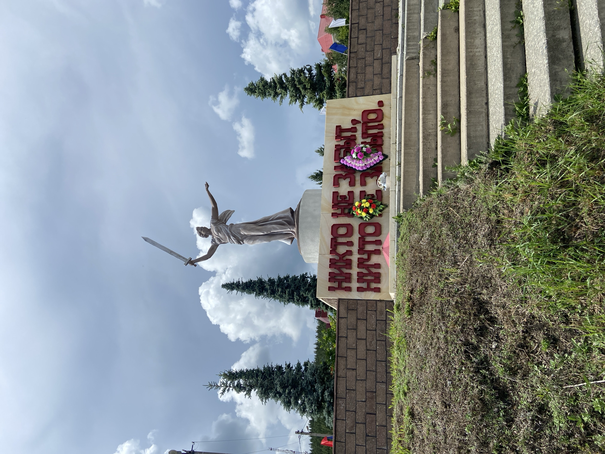 In the city of Askarovo, Russia, a memorial monument stands against a cloudy sky. The monument features a statue of a woman holding a sword aloft. In front of the statue, red text is inscribed on a beige background. Two wreaths, one with yellow and red flowers and another with pink and purple flowers, are placed at the base of the monument. Concrete stairs lead up to the monument, and the surrounding area is covered in grass. Brick walls and several evergreen trees are visible behind the monument.