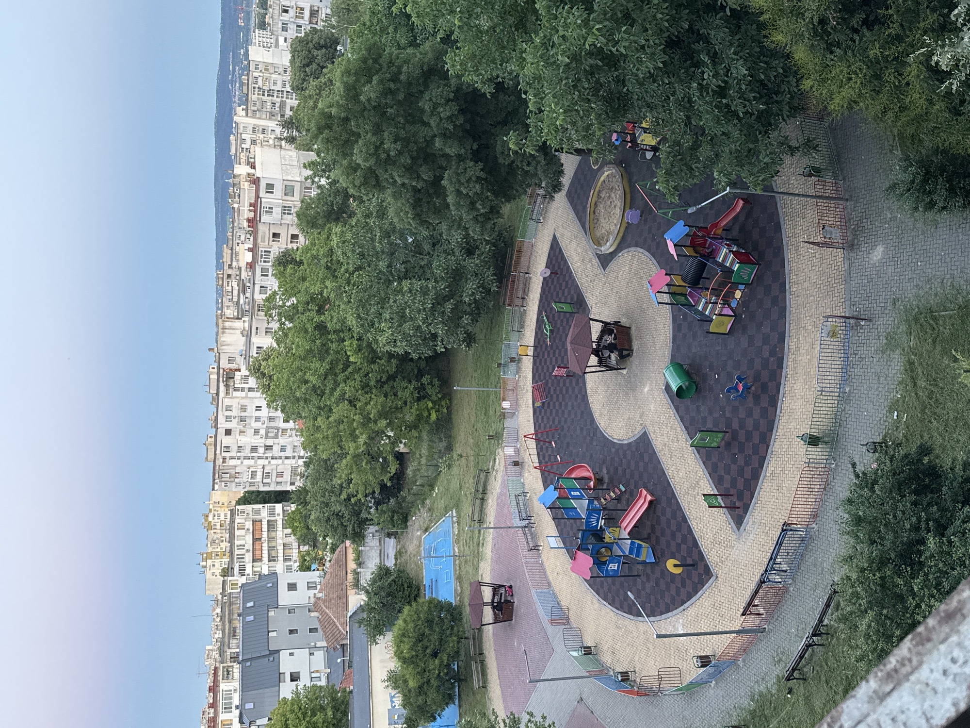 The image depicts a children's playground in the city of Varna, Bulgaria, located in a residential area surrounded by apartment buildings. The playground features various play structures, including slides, swings, a climbing frame, and a sandbox. The ground is covered with a black rubber safety surface, and the pathways are paved with brick. A small, hexagonal gazebo with a brown roof is situated in one corner of the playground, providing shade and a place to sit. Several benches are placed around the playground area for seating. In the background, numerous multi-story apartment buildings are visible, typical of urban European landscapes. Lush green trees are interspersed throughout the scene, softening the urban environment. The sky is a pale blue, suggesting either early morning or late evening light. There are no visible people actively engaged in play within the main playground area, though two individuals are seated inside the gazebo. The overall scene presents a well-maintained community space within a densely populated urban setting.