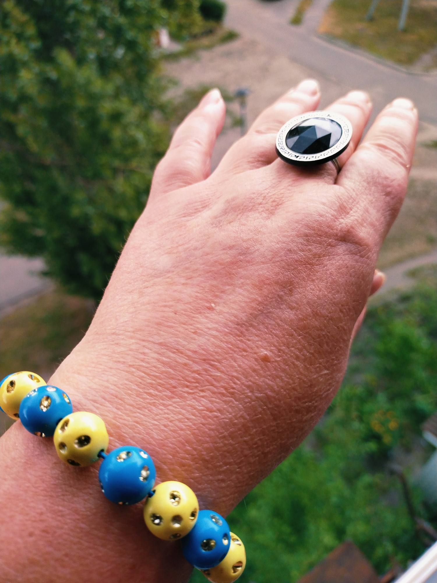 The image shows a close-up of a person's hand wearing a ring and a bracelet. The ring has a large, faceted black stone set in a silver-toned band with decorative etching. The bracelet is made of alternating blue and yellow beads, each with multiple small holes and what appear to be metallic accents within the holes. The background is blurred, showing greenery and a hint of a street or pathway, suggesting an outdoor setting. The context of Horishni Plavni, Ukraine, is external information not directly discernible from the visual content of the image itself.