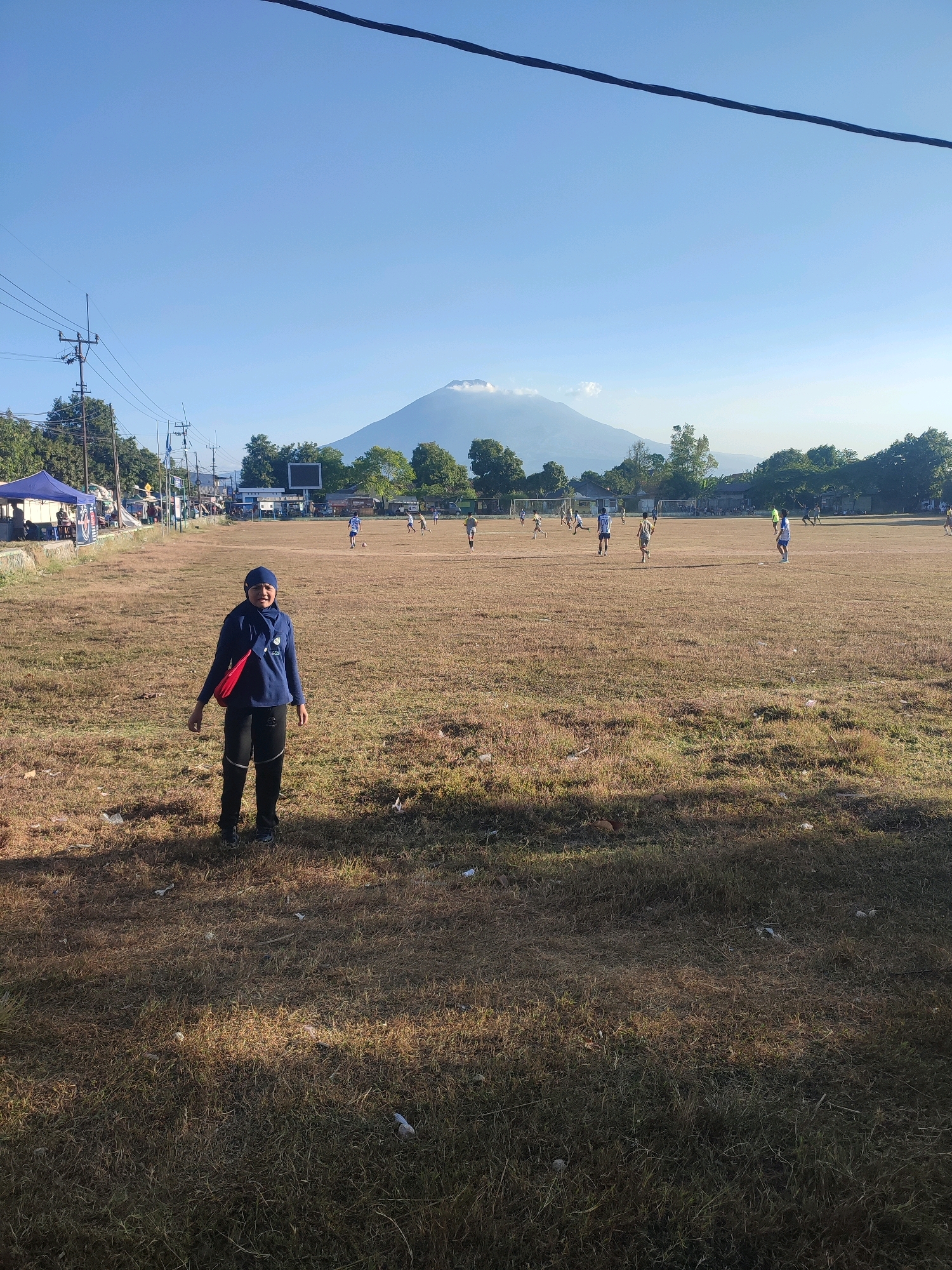 The image depicts a person standing on a dry, grassy field, likely a sports ground, with a soccer game in progress in the background. A large mountain dominates the horizon, with a plume of smoke or clouds near its summit, suggesting it might be an active volcano. The sky is clear and blue. In the distance, buildings and trees are visible, along with power lines and utility poles. There are tents and what appear to be spectators or participants gathered along the side of the field. The ground is covered in dry grass and scattered litter. The overall scene suggests an outdoor event taking place in a rural or semi-urban area. The location is Tegalwangi, Indonesia.