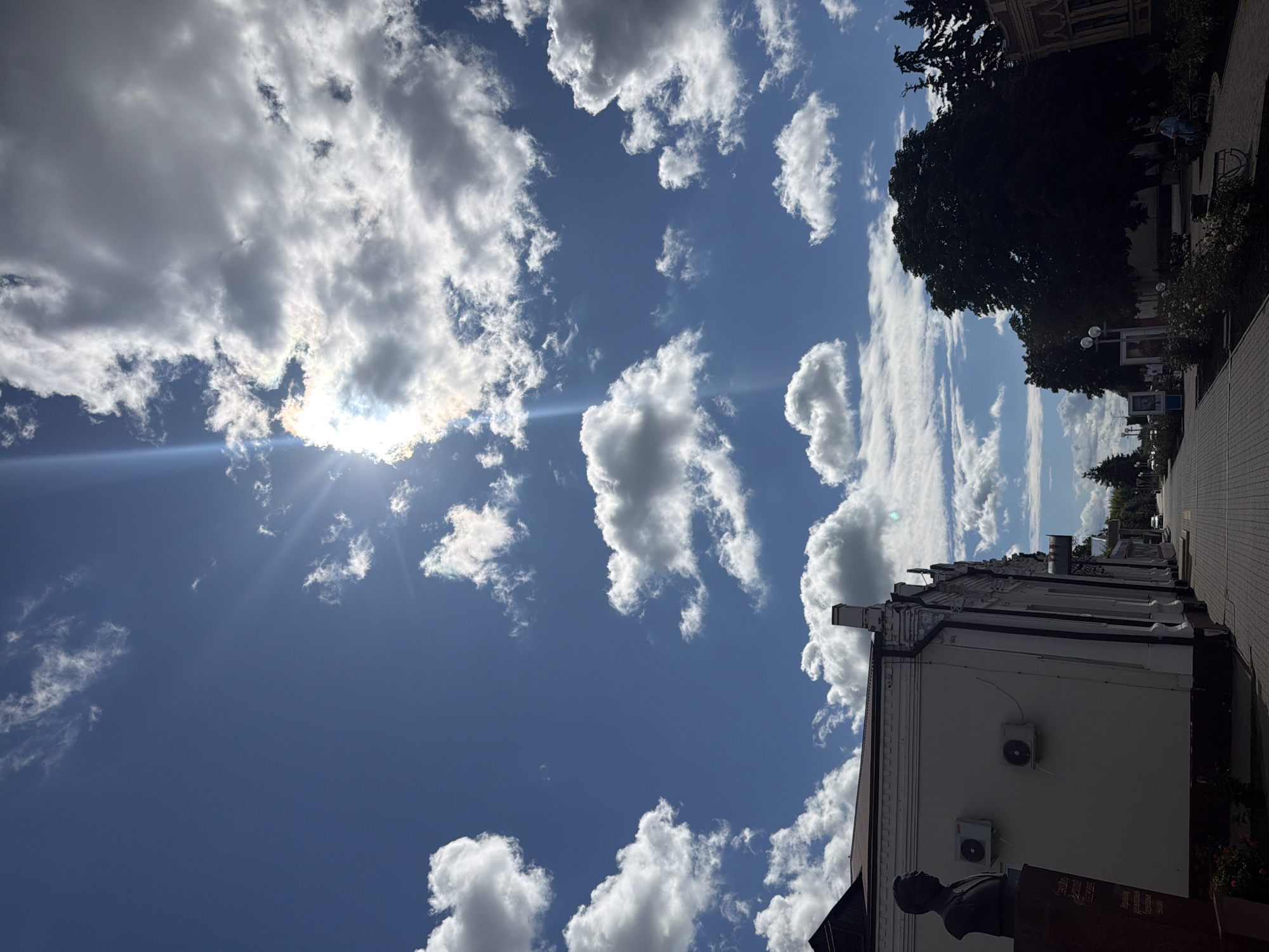 The image presents a scene in Leningradskaya, Russia. The sky is predominantly blue with scattered cumulus clouds, and the sun is visible through a cloud layer, creating a bright, diffused light. The right side of the frame includes part of a building facade, likely made of painted stucco, with visible architectural details. An air conditioning unit and a bust monument are visible against the building. A tree line is partially visible above the buildings in the background. A paved sidewalk is on the right side of the image. Several figures walk towards the horizon on the sidewalk.