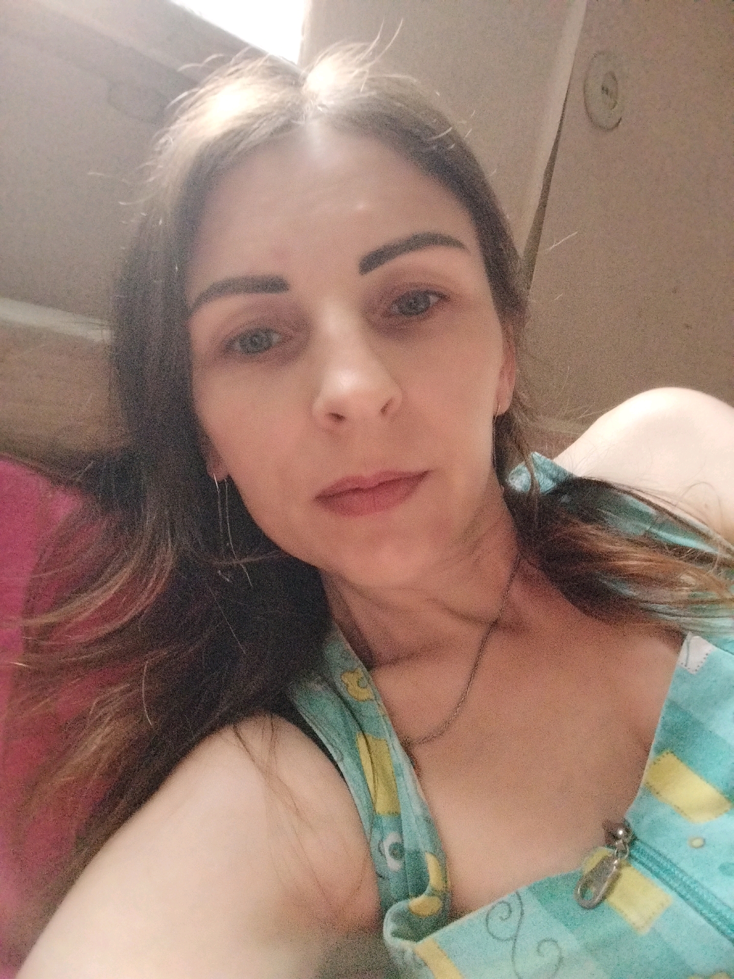 The image is a close-up, selfie-style photograph of a woman. She has long, dark brown hair that is slightly wavy and falls over her shoulders. Her eyes are a light color, and she is looking directly at the camera. She is wearing a turquoise and yellow patterned top with a zipper in the front. A thin silver chain necklace is visible around her neck. Her makeup appears to include eyeliner and possibly eyeshadow. The background is indistinct, with hints of a textured wall and a bright light source, possibly from a window. There are no specific landmarks or environmental cues to suggest a location within Vinnytsia, Ukraine.