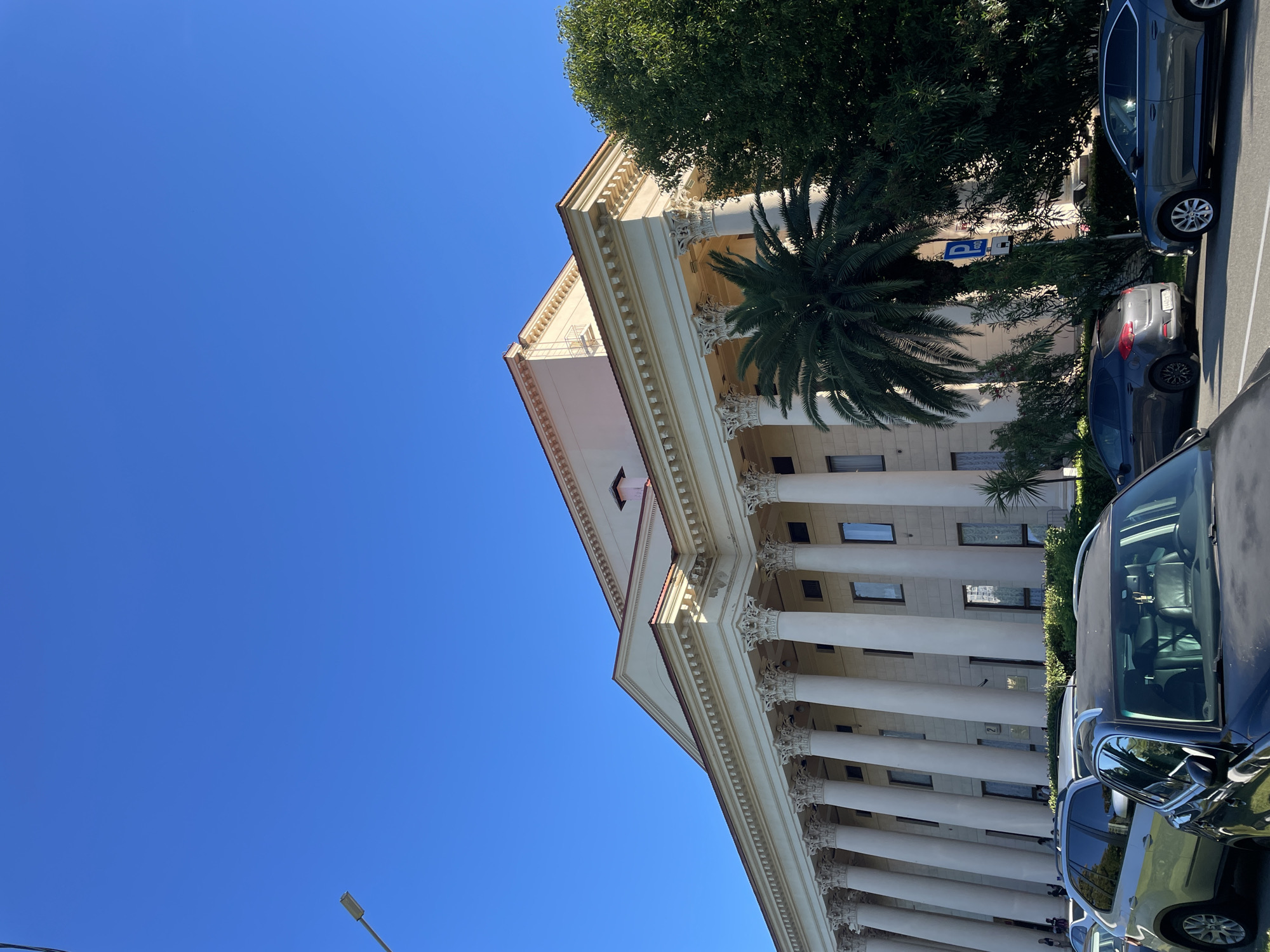 The image shows a large, classical-style building with a colonnade of tall, white pillars under a clear blue sky. The building appears to be the Winter Theater in Sochi, Russia, as suggested by its architectural features and the presence of palm trees, which are characteristic of the region. In the foreground, several cars are parked on a street in front of the building, indicating a public space. The scene is well-lit, suggesting it was taken during daylight.