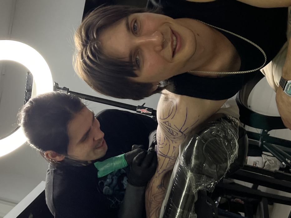 An indoor scene in a tattoo studio in Rostov-on-Don, Russia, depicts a tattoo artist working on a client. The tattoo artist, a male with dark hair pulled back and multiple ear piercings, wears a black t-shirt and black gloves, leaning over to meticulously apply a tattoo to the client's left arm using a machine covered in a green disposable barrier. He displays a subtle smile and focused expression. The client, a male with medium-length brown hair, wearing a black tank top, a silver chain necklace, and a silver septum piercing, is positioned in the foreground, smiling directly towards the camera. Purple stencil lines for a complex design are visible on his left arm, which rests on a plastic-wrapped padded support. A large, illuminated ring light on a stand provides primary illumination from the background. A black stand with a red star sticker is also visible. The client wears a digital watch on his right wrist. The background consists of plain, light-colored walls.