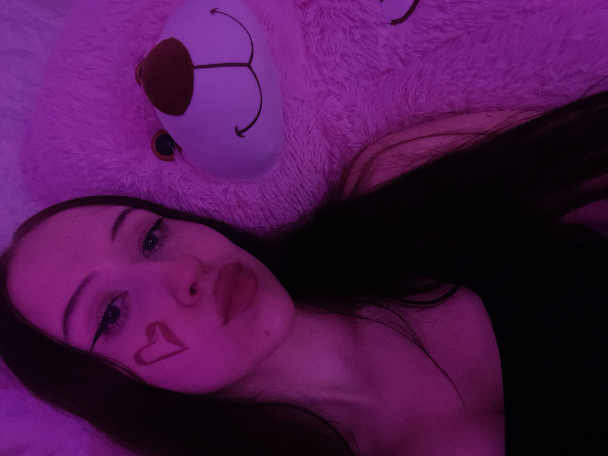 A young woman with dark hair and a heart drawn on her cheek is posing for a selfie with a large pink teddy bear. The lighting is pink, creating a vibrant and intimate atmosphere. She is wearing a black top. The teddy bear is fluffy and has a stitched smile. The scene appears to be indoors, possibly a bedroom, with a soft, plush surface suggesting bedding or a rug. No specific location in Votkinsk, Russia, can be determined from the image.