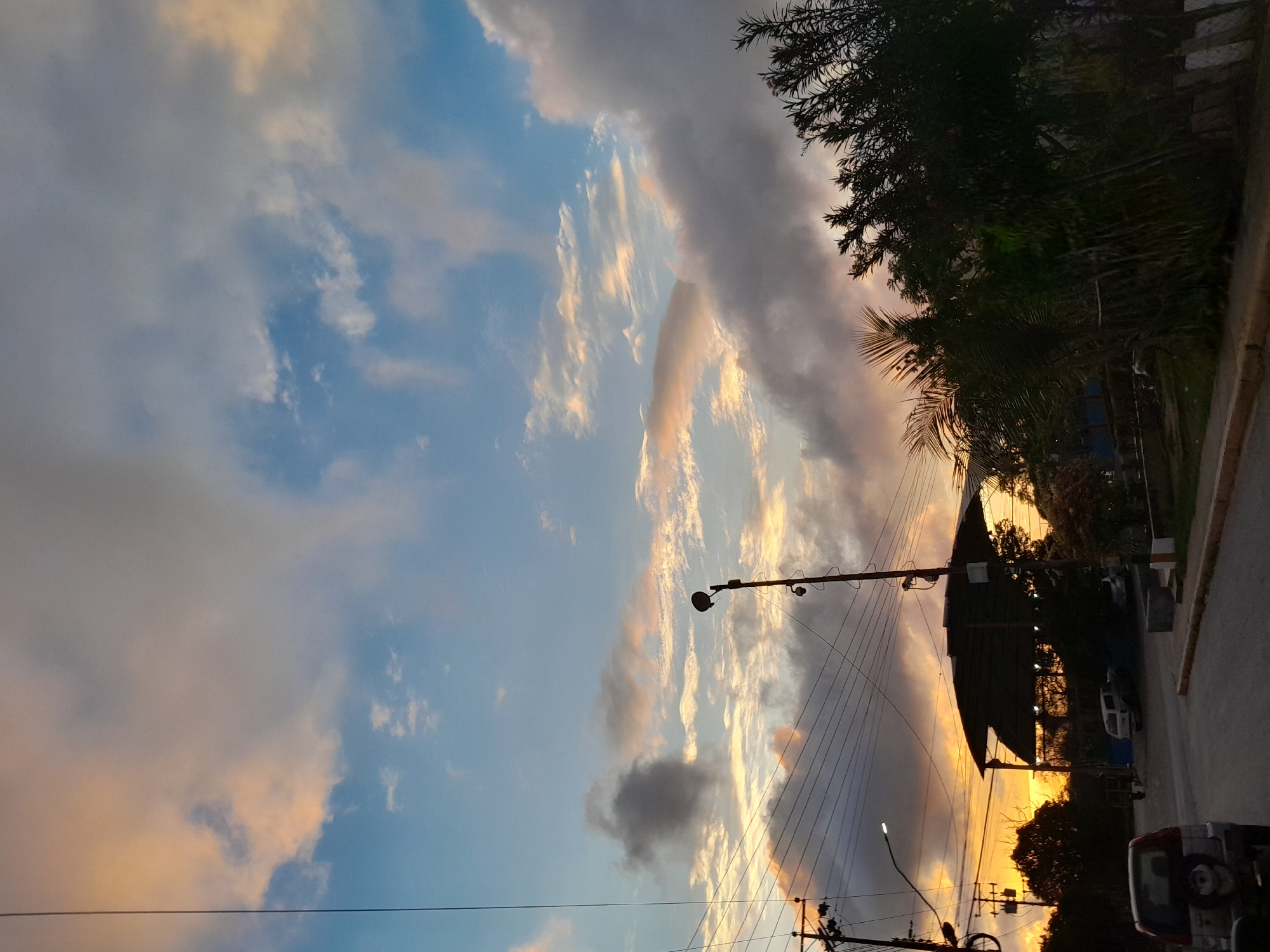 The image captures a twilight scene on a street in Santa Ana, Venezuela. The sky is a dramatic canvas of orange, pink, and blue hues, painted by the setting sun behind a cluster of clouds. A street lamp on a pole stands prominently in the middle ground, casting its light downwards. Power lines crisscross the sky, creating a complex network above a covered structure that appears to be a carport or open-air shed.
In the foreground, the rear of a silver SUV is visible on the asphalt road. Further down the road, other vehicles, including a white SUV, are parked or driving. Dense foliage and trees line the right side of the street, with some palm fronds peeking out. The overall impression is one of a quiet, semi-urban environment at the end of the day, with a focus on the atmospheric sky and the infrastructure of the street. There are no people visible in the scene.