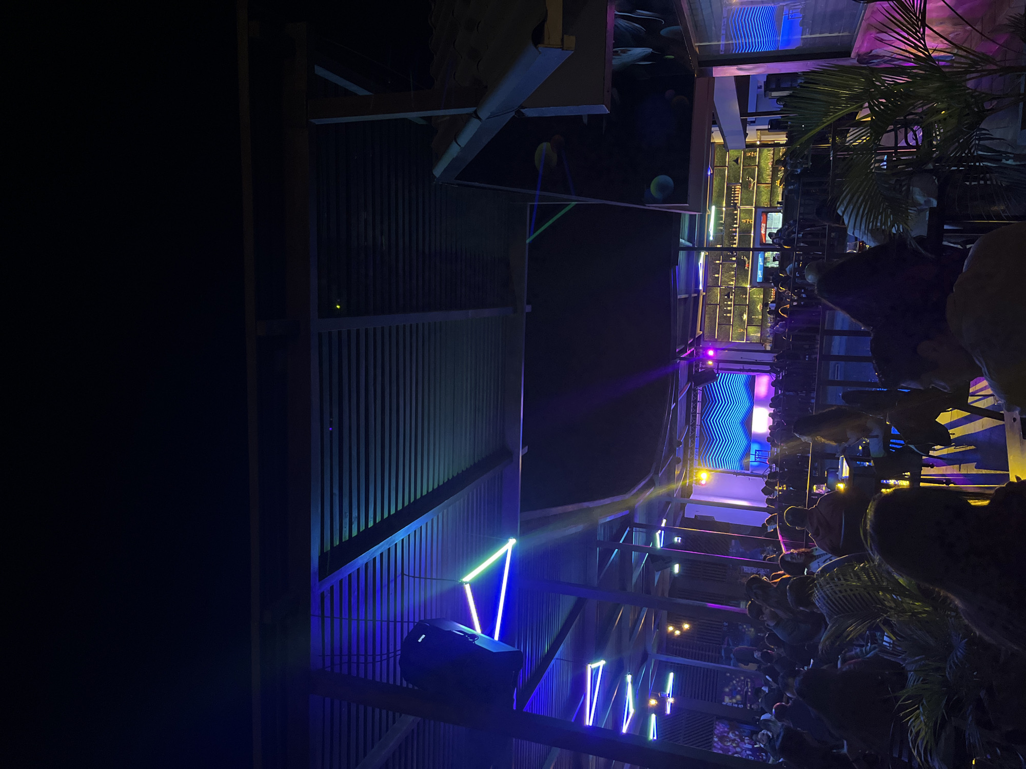 The image shows a nighttime scene at a bar in Arica, Chile. Patrons sit at tables and stand on a tiered patio. A stage with a screen displaying an abstract, wavy blue graphic is visible in the background. The venue is decorated with linear light fixtures in blue and green and some plants. A large speaker is mounted on the structure. The patio is made of metal latticework. Some of the surfaces have a painted mural.
