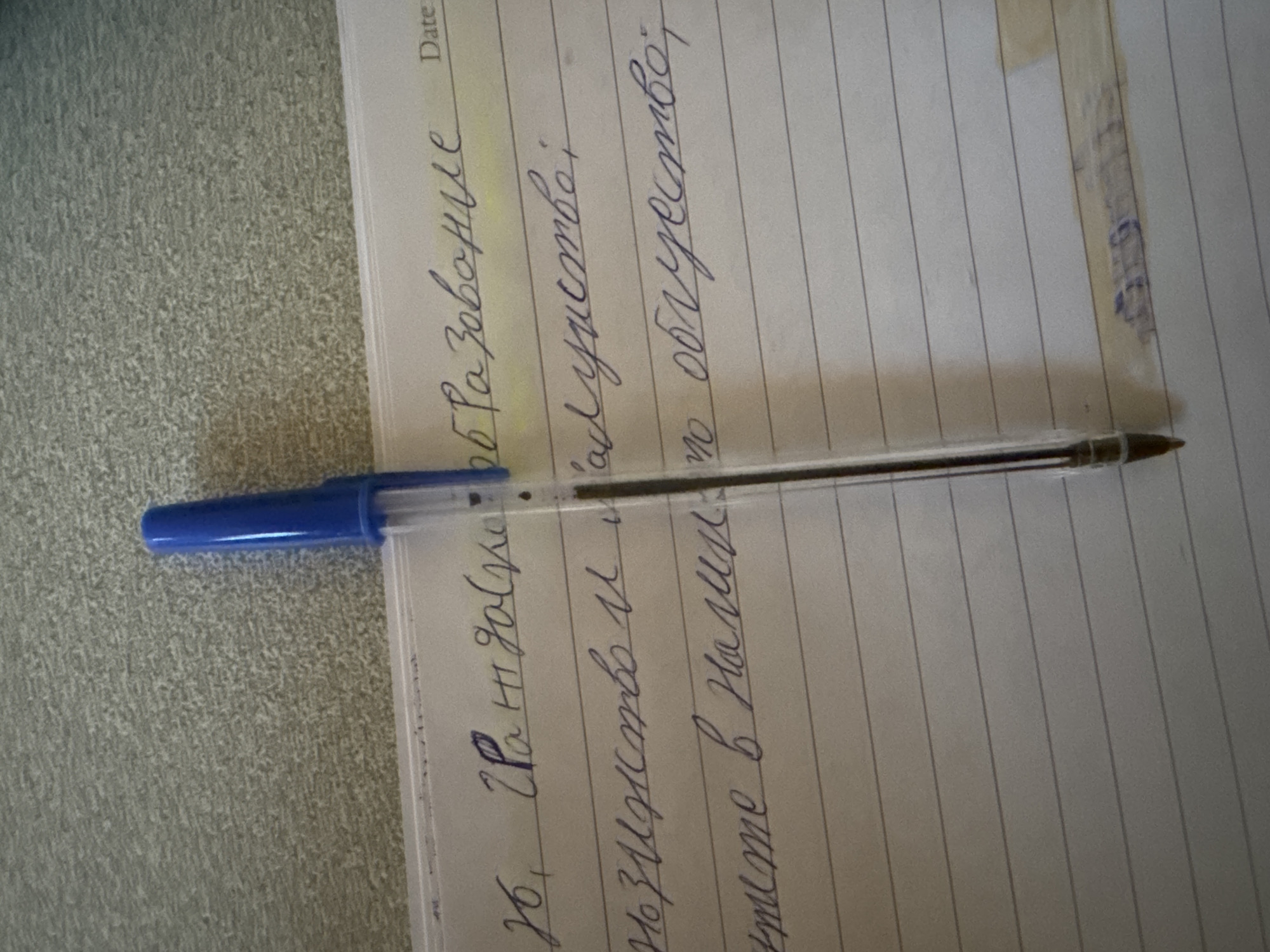 A close-up shows a lined sheet of paper with handwritten text in blue ink, oriented horizontally. The text includes the words "образование" and "облусство." A blue ballpoint pen lies across the page, its tip pointing towards the right. The paper rests on a surface with a light gray, textured pattern. A narrow strip of adhesive tape is affixed to the top right corner of the paper. The text on the paper appears to be in Russian. There is no information related to Burgas, Bulgaria.