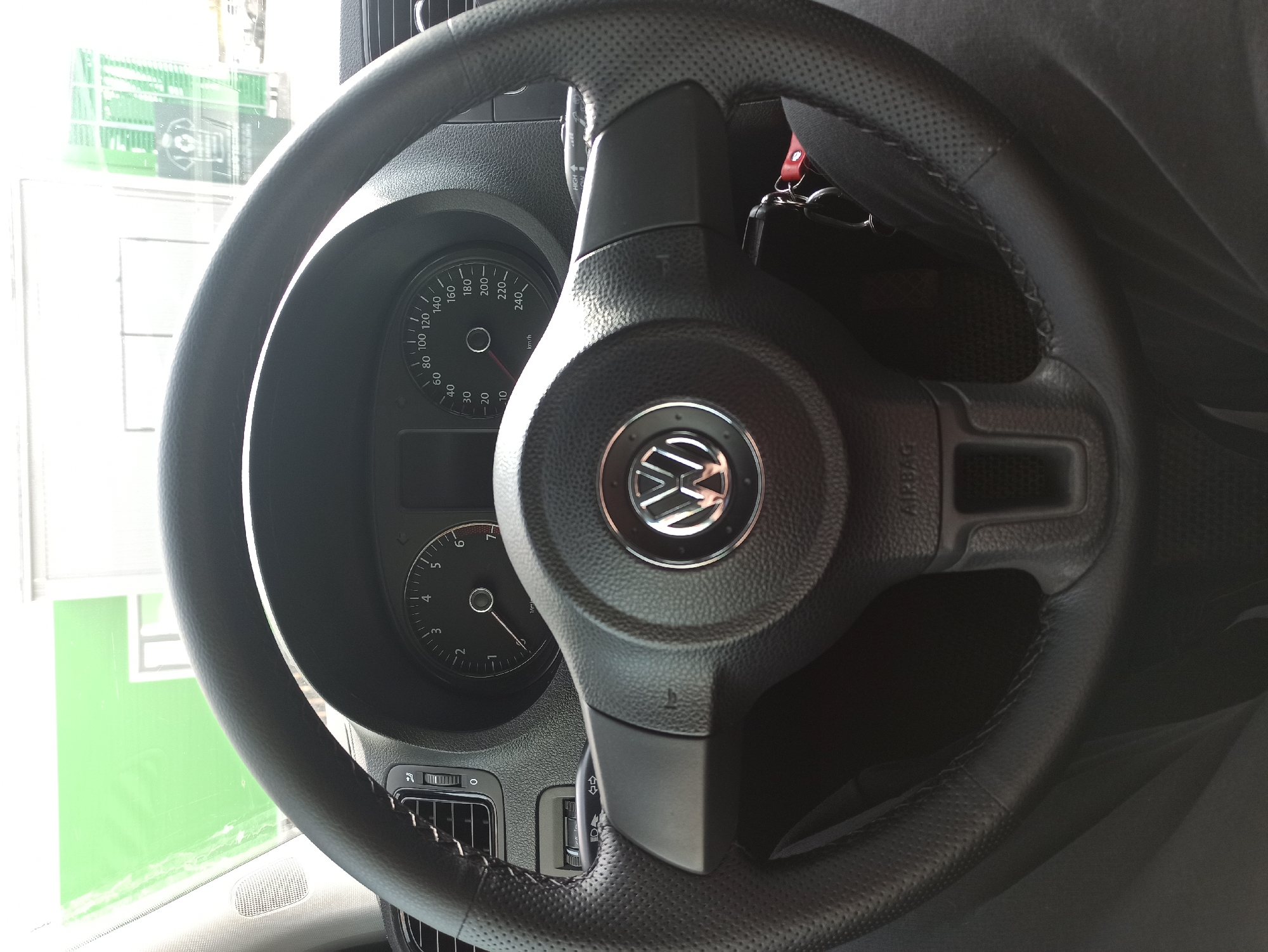Here is a detailed description of the image:
The image shows the steering wheel and dashboard of a Volkswagen vehicle. The steering wheel is black leather with a prominent Volkswagen logo in the center. Behind the steering wheel, the instrument panel is visible, featuring a speedometer, a tachometer, and other gauges. The speedometer displays markings in kilometers per hour, ranging from 0 to 240 km/h. A label that says "AIRBAG" is visible on the right spoke of the steering wheel.
A window can be seen to the left of the steering wheel, showing an out-of-focus view of what appears to be a brightly lit interior space with white and green elements. Based on the provided information, no specific location in the city of Unknown, Unknown, can be determined from this image. No people, subjects, interactions, or actions are visible in the image.