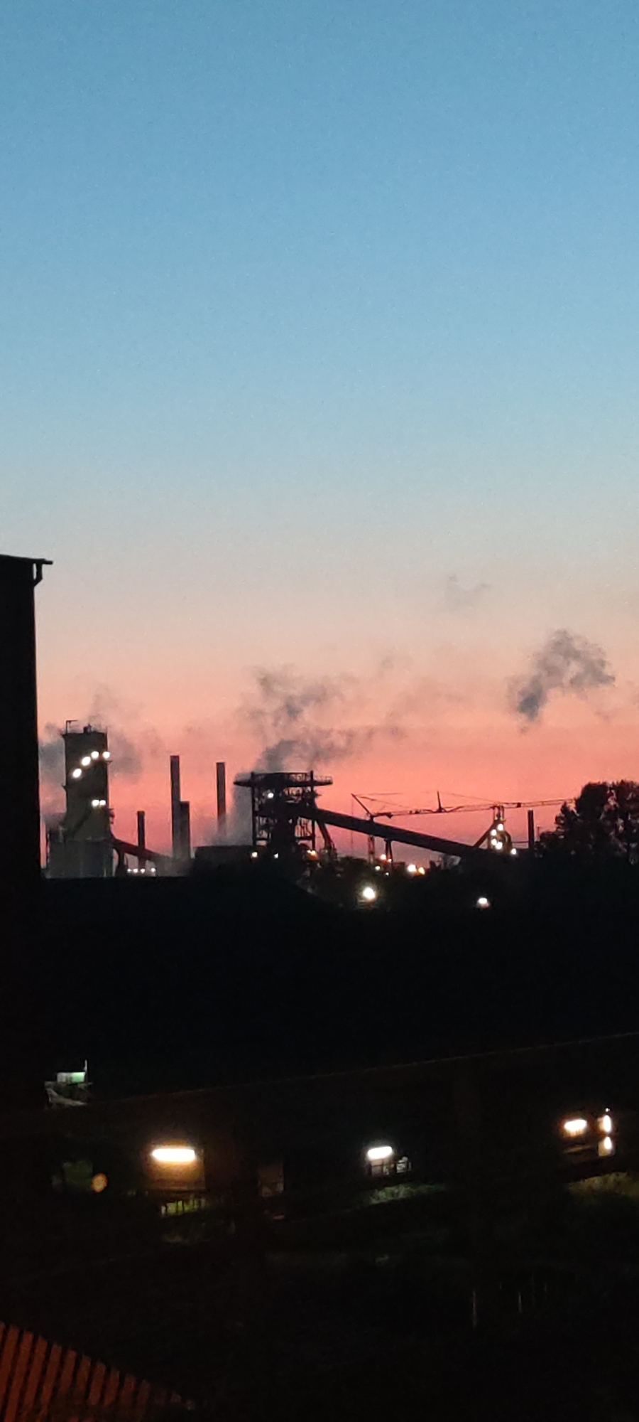 The image depicts an industrial landscape at dusk, likely in Salzgitter, Germany, given the context of industrial facilities. The sky transitions from a clear blue at the top to a soft pink and orange gradient near the horizon, indicating sunset or sunrise.
Prominently featured in the mid-ground are structures of a steelworks or similar industrial plant. Tall, silhouetted chimneys and towers rise against the colored sky. Smoke or steam is visibly emanating from some of these structures, creating a hazy effect. Various platforms, conveyor belts, and cranes are discernible, forming the complex architecture of the industrial site. Artificial lights are visible across the structures, some shining brightly, others appearing as scattered points of illumination.
In the foreground, silhouetted against the industrial scene, is a dark area that appears to be a collection of buildings or structures, possibly residential or other urban elements. Some rectangular lights, possibly from windows or streetlights, are visible within this foreground area, suggesting a human presence or activity despite the dim lighting. The very bottom of the image shows a dark, textured surface that might be a fence or railing. The overall impression is one of an industrial city at the transition between day and night, with human activity juxtaposed against a large-scale industrial environment.