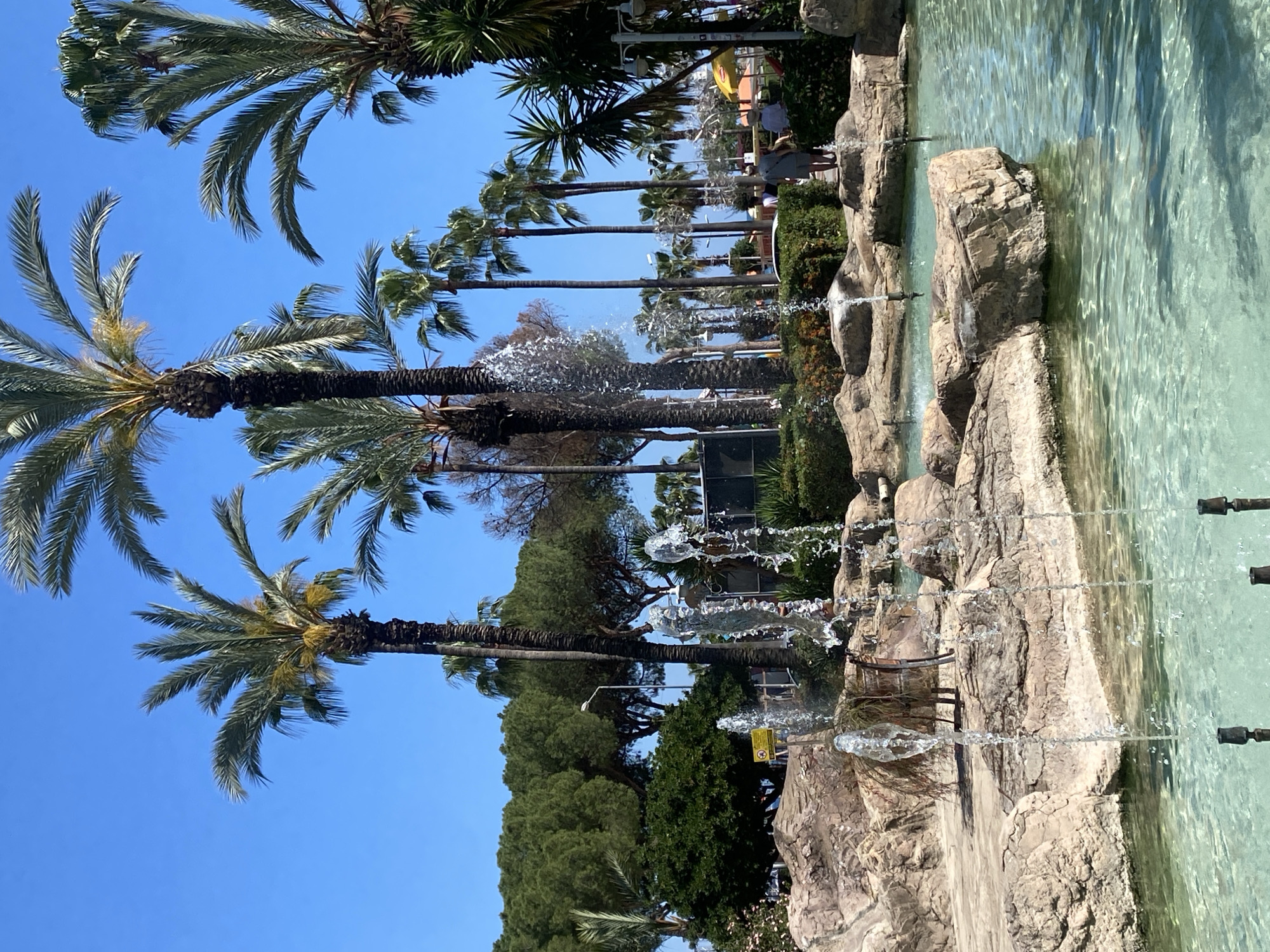 The image depicts a fountain with several streams of water cascading into a pool. The fountain is constructed with rocks, and the water appears clear and turquoise. In the background, there are palm trees against a clear blue sky. Further in the background, people can be seen walking in the distance. The image is likely taken in the city of Alanya, Turkey.