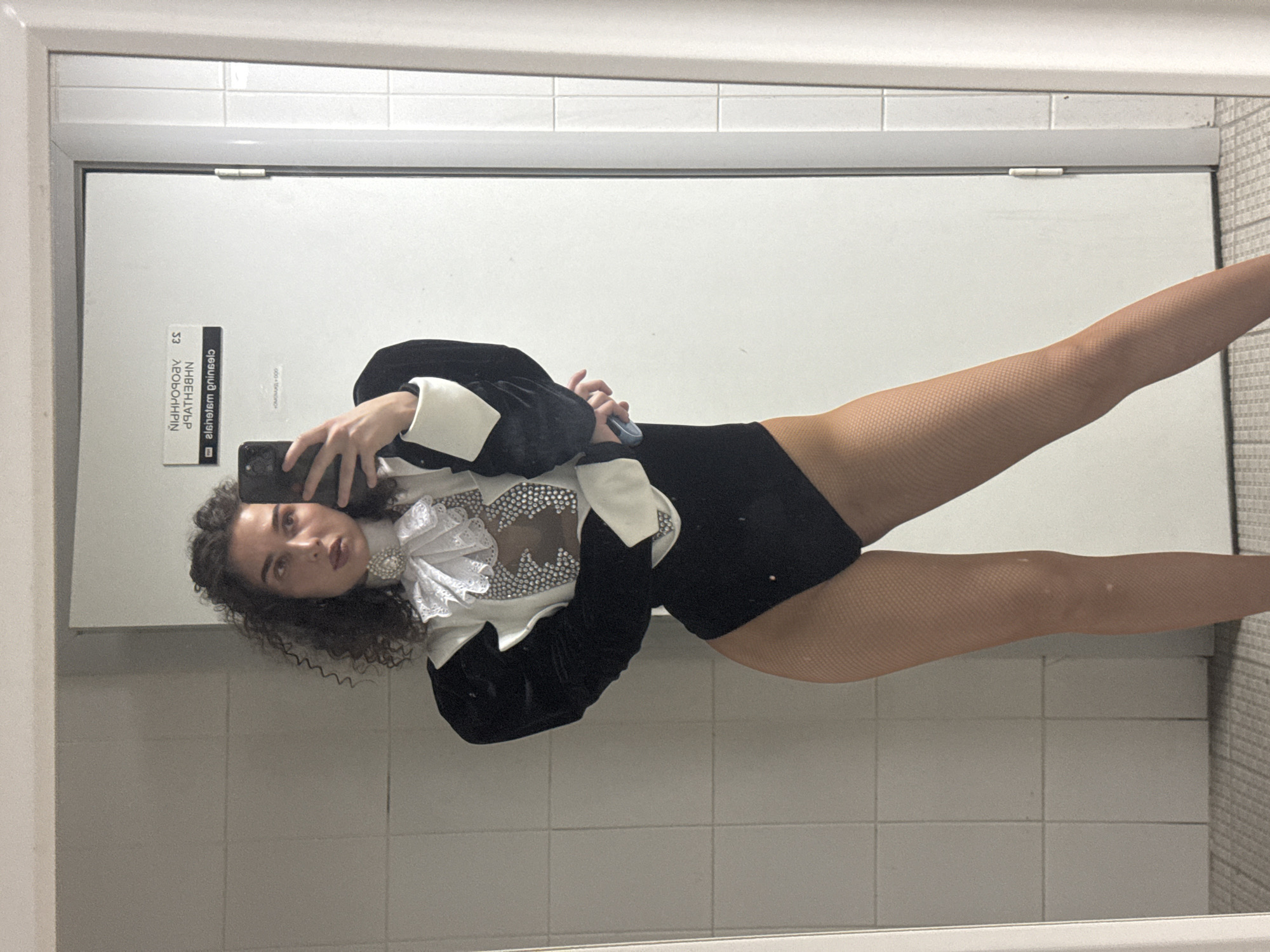 A woman is taking a mirror selfie in what appears to be a locker room or bathroom. She is wearing a black leotard with a white and black ruffled collar, adorned with rhinestones. The leotard has voluminous puffed sleeves with white cuffs. She is also wearing fishnet stockings. The background consists of tiled walls and a white door with a sign. The sign on the door contains text that appears to be in Cyrillic alphabet, which suggests a location in Russia. The text on the sign is reversed due to the mirror reflection. The text "NÏDHPO90AV ES 9ATH3BHN" and "algihelsm gninsels" are visible. These inscriptions, combined with the presence of Cyrillic, strongly indicate the location is in the city of Kazan, Russia. The woman is holding a smartphone in her right hand, capturing her reflection.