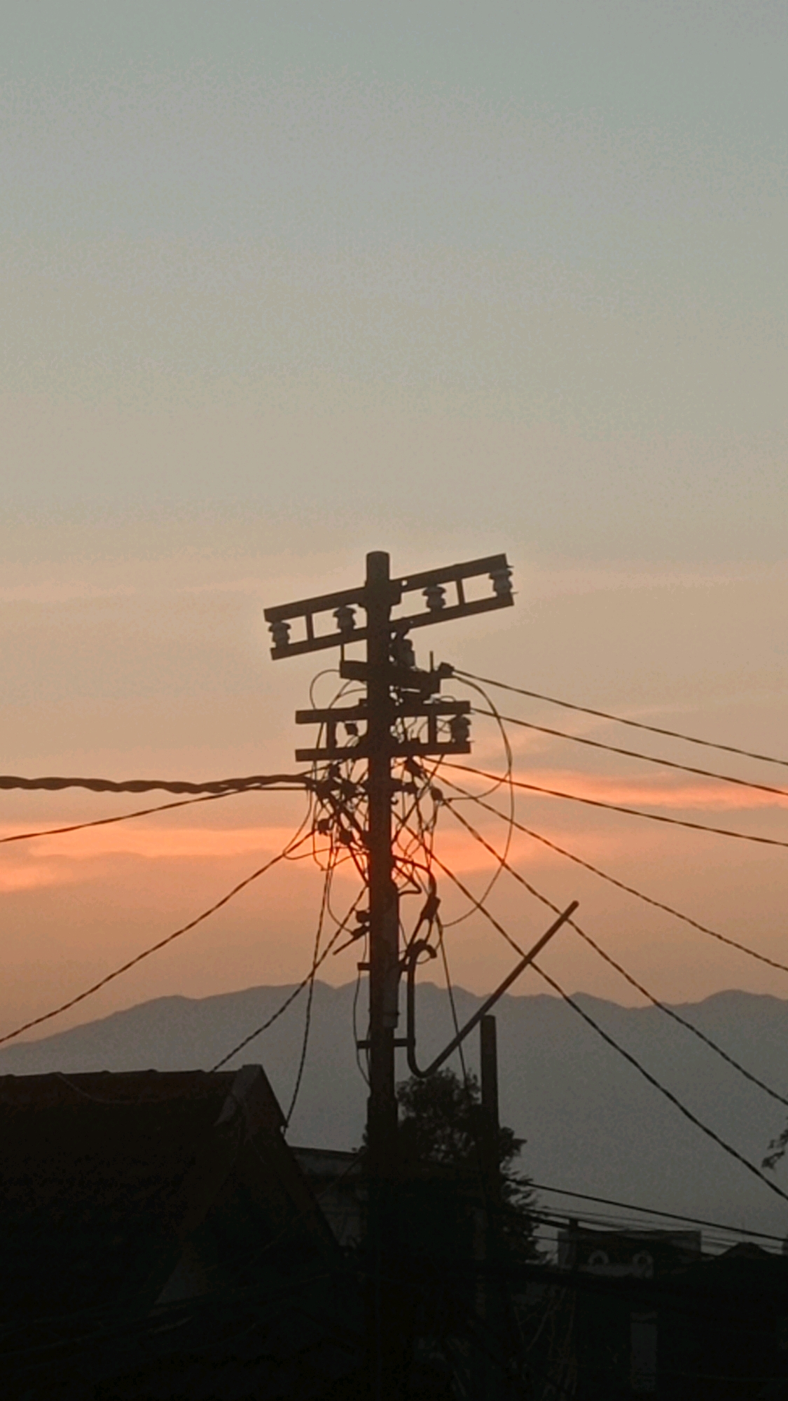 A vertical shot captures a power pole silhouetted against a hazy, orange-tinted sky at dusk. The pole is laden with a dense network of tangled electrical wires and insulators. In the foreground, the dark silhouette of a rooftop is visible, and a range of layered mountains forms a backdrop. The overall scene is tinged with the muted colors of sunset, with a gradient of pale blue to soft orange dominating the sky. The location is the city of Mulyoagung, Indonesia.