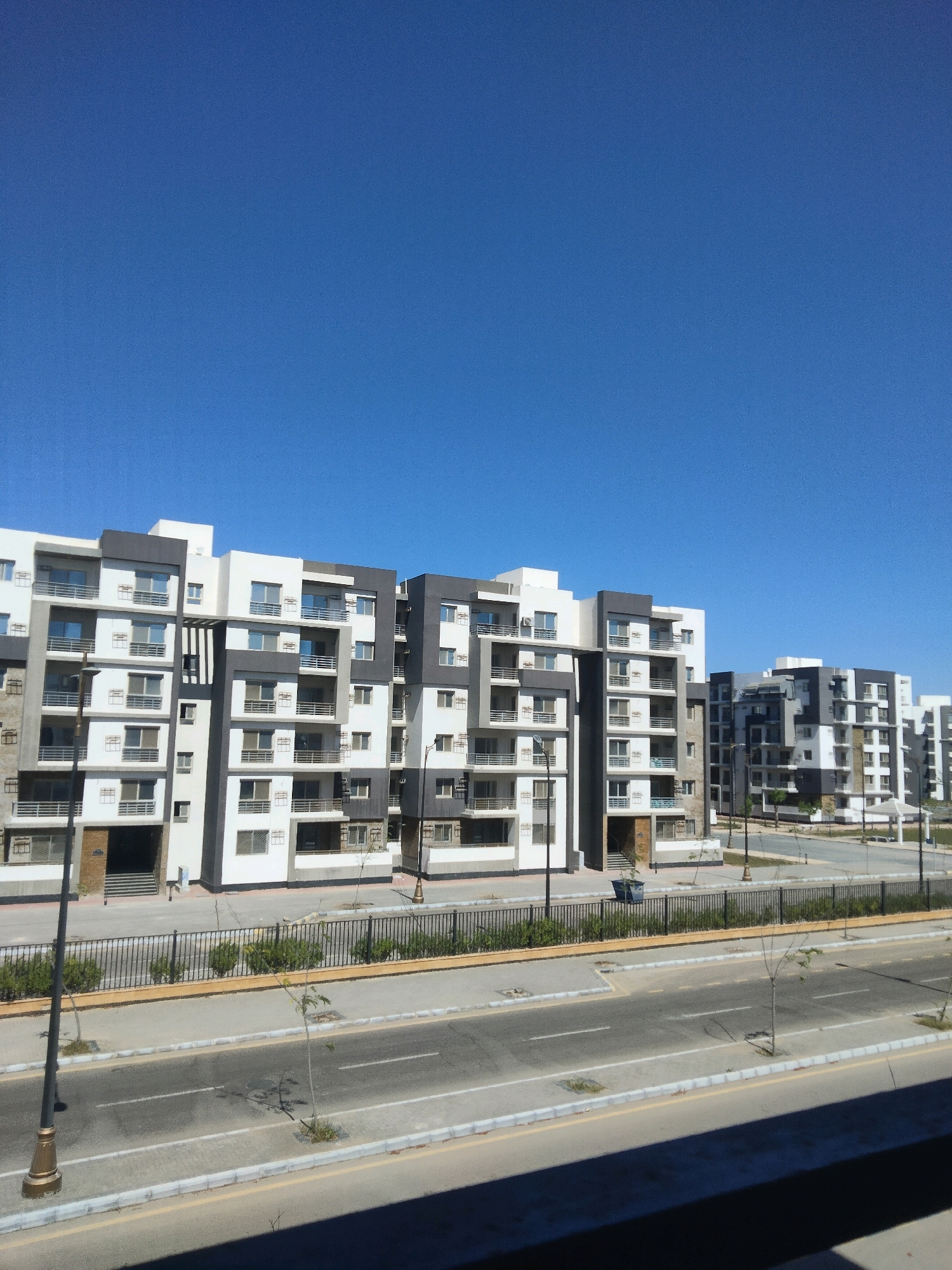 The image shows a modern residential complex in the city of Gamasa, Egypt, under a clear blue sky. The buildings are multi-story apartment blocks with a contemporary design, featuring a combination of white and dark gray facades. Balconies with metal railings are visible on multiple floors. In front of the buildings, there is a paved road with visible lane markings. A black metal fence runs along the perimeter of the complex, separating it from the road. Small trees and bushes are planted along the road and within the complex. Streetlights are also present. The overall scene depicts a quiet, urban residential area.