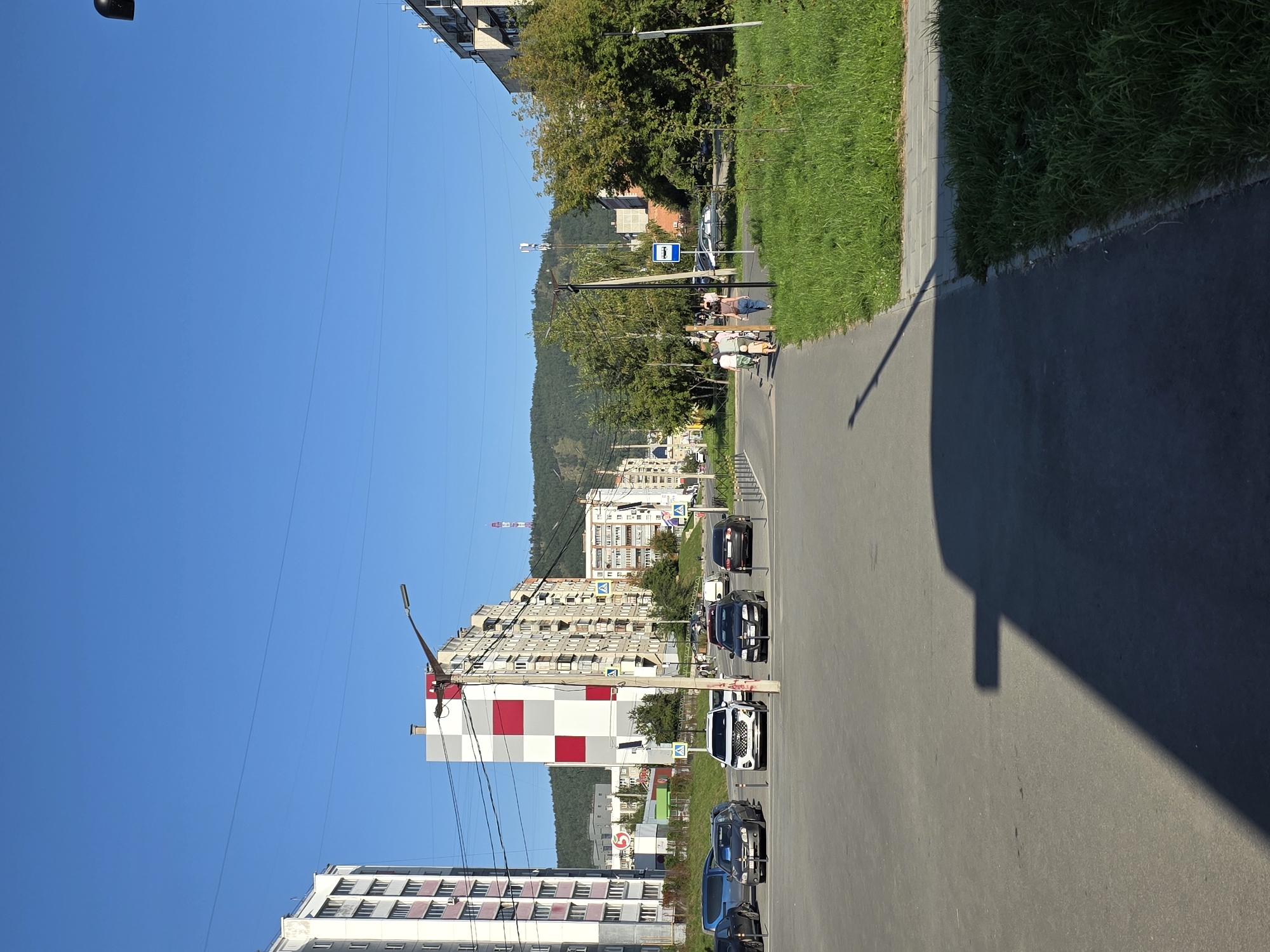 This image depicts a street scene in the city of Miass, Russia, on a clear, sunny day. The foreground shows a paved road with a sidewalk and a patch of lush green grass to the right. A long shadow of an unseen object, possibly a building or a tall structure, stretches across the road from the left side of the frame. Several cars are parked or driving on the road, including a white SUV and several sedans. A bus stop sign is visible on the right side of the road, adjacent to the sidewalk.
In the mid-ground, apartment buildings with multiple stories line the street. One prominent building is characterized by a distinctive checkered facade of red and white squares. Another taller building on the left is white with pink accents. Trees are interspersed between the buildings and along the roadside.
The background features a range of hills or mountains, covered in green vegetation, under a bright blue sky. A tall, slender tower, possibly a communication tower or a radio mast, is visible on the distant hills. Several people are walking on the sidewalk in the mid-ground, appearing to be engaged in casual conversation. The overall impression is of a residential area in a city with a backdrop of natural landscape.