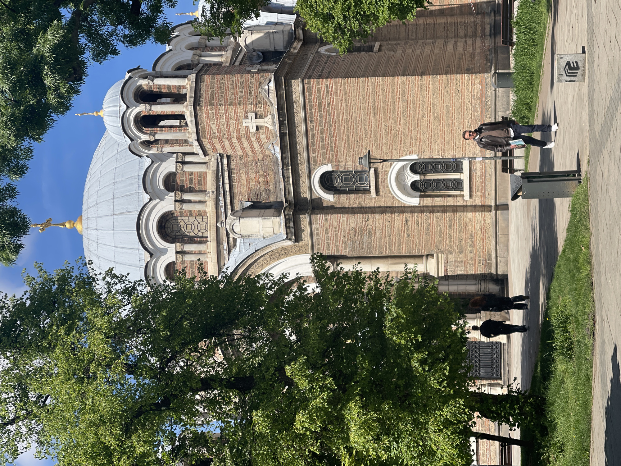 The image shows a building in Sofia, Bulgaria, most likely the Alexander Nevsky Cathedral, partially obscured by trees. The building is brick and stone, with arched windows and a dome. A man in a leather jacket, jeans, and white sneakers stands on the sidewalk next to a bench and a trash can; he is holding papers. Two more people are walking away from the building, they are indistinct. The sky is blue and clear. The environment suggests an urban park setting.
