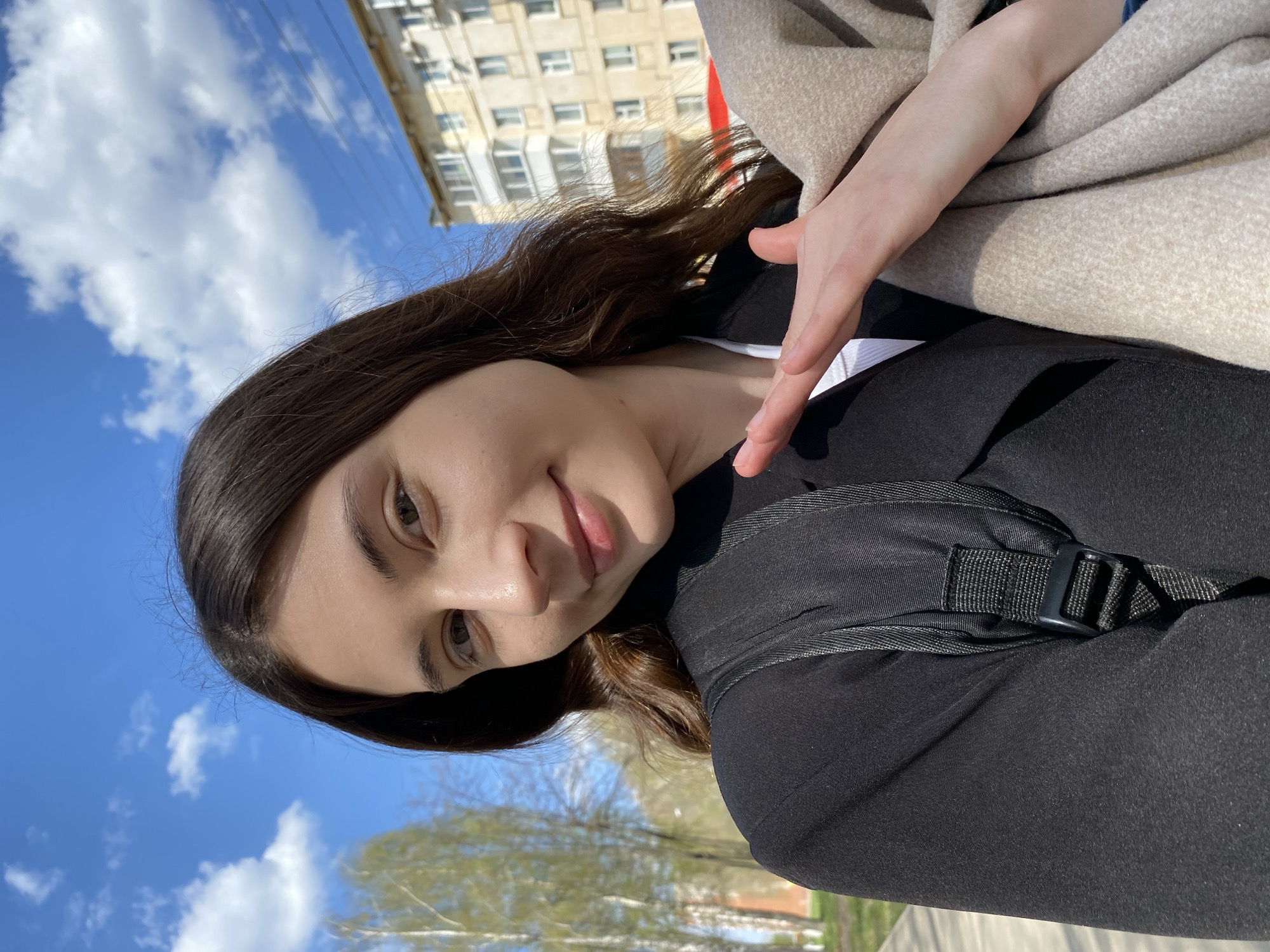 A woman poses for a selfie on a sunny day in Votkinsk, Russia. She has long, dark hair, fair skin, and wears a black jacket with a visible backpack strap. A beige coat is draped over her shoulder. In the background, a multi-story building with white facades and balconies is visible, along with a blue sky dotted with fluffy white clouds. A row of green trees lines a street, adding to the urban landscape.