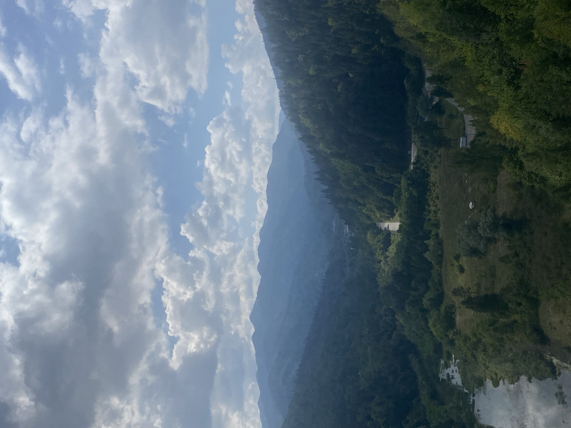 This is an aerial, wide shot of a mountainous landscape. The scene is dominated by a valley covered in lush green trees, with a winding road visible through the foliage. A river flows along the left side of the frame, reflecting the sky and clouds. In the background, several layers of misty blue mountains recede into the distance, creating a sense of depth. The sky above is partly cloudy, with patches of blue sky visible between the white, puffy clouds. The overall impression is one of natural beauty and serenity, suggesting a location within the Carpathian Mountains near Bicaz, Romania, a region known for its dramatic topography and dense forests.