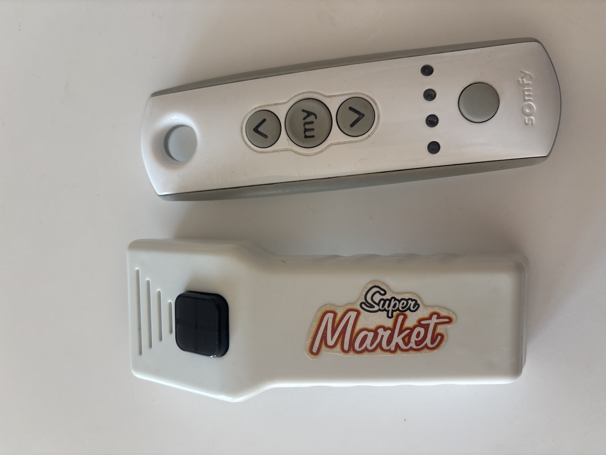 Two remote controls are placed on a white surface. The remote on the left is white and elongated, with a black button at the top and a sticker with "Super Market" written in red and white. The remote on the right is also white and elongated, with a silver-grey colored side. It features three circular buttons arranged vertically: an upward-pointing arrow, a button with "my" inscribed on it, and a downward-pointing arrow. Below these, there are four small circular indicators and a larger circular button. The word "somfy" is embossed at the bottom of this remote. The image is taken from a top-down perspective, and the lighting suggests an indoor environment. There are no people or animals visible in the image. The setting does not contain any specific landmarks or contextual elements to determine its location within Dubai, United Arab Emirates.