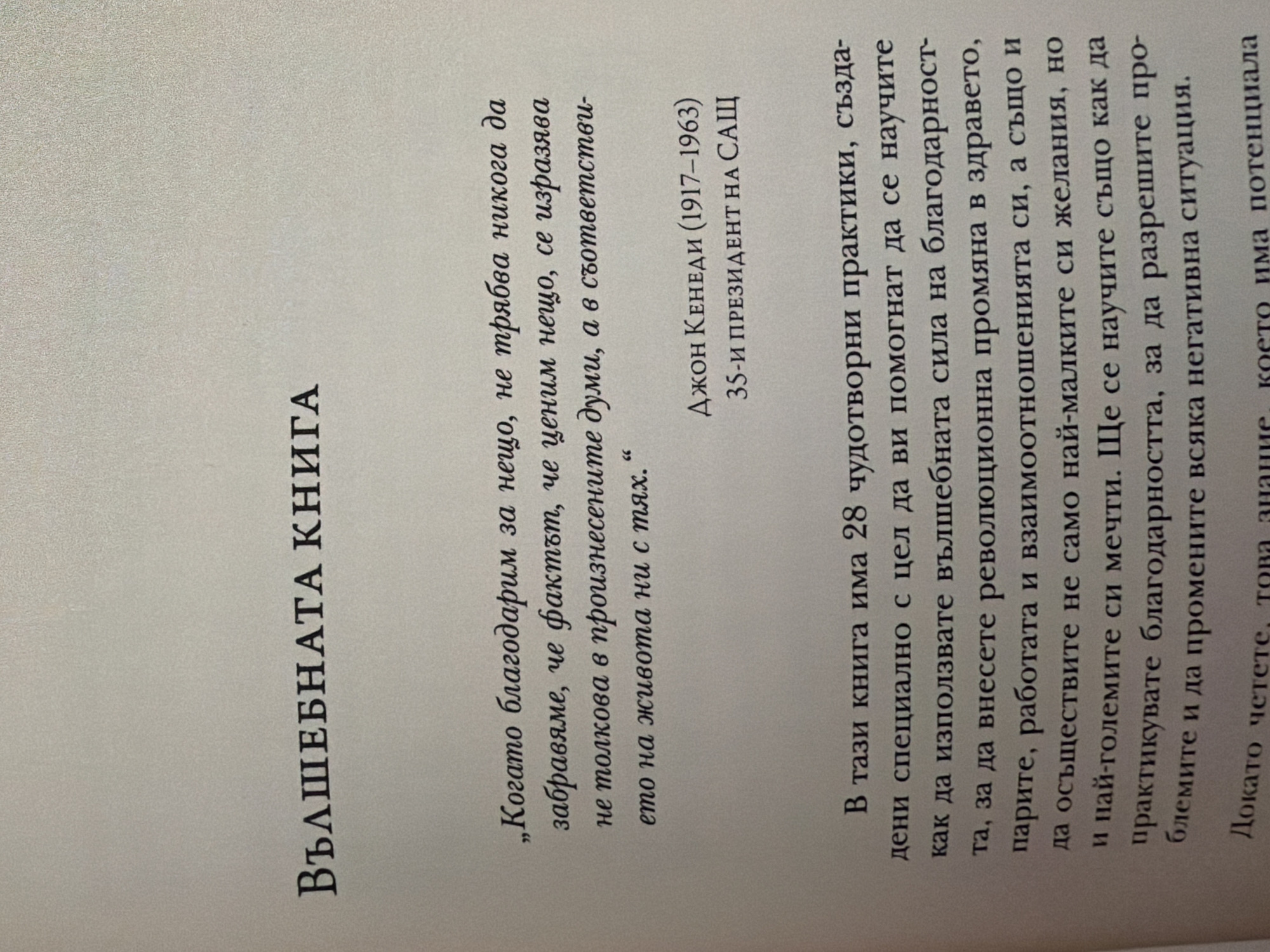 The image contains a close-up, angled shot of a page from a book. The text is in Bulgarian and appears to be a quote and introductory text.
The quote, attributed to John F. Kennedy (1917-1963), 35th President of the USA, reads: "When we give thanks for something, we must never forget that to value something is expressed not so much in spoken words, but in actions in life with them."
Below the quote is introductory text that begins: "This book contains 28 miraculous practices that will help you learn how to use the power of gratitude to bring about revolutionary change in your health, relationships, work and subconscious, as well as to fulfill not only your smallest but also your biggest dreams. You will also learn how to practice gratitude, to allow yourself to overcome any negative situation."
There are no identifiable people, actions, or interactions visible in the image. The environment is a plain, off-white surface, consistent with a book page. The location is not determinable from the image itself, and therefore cannot be associated with Pazardzhik, Bulgaria.