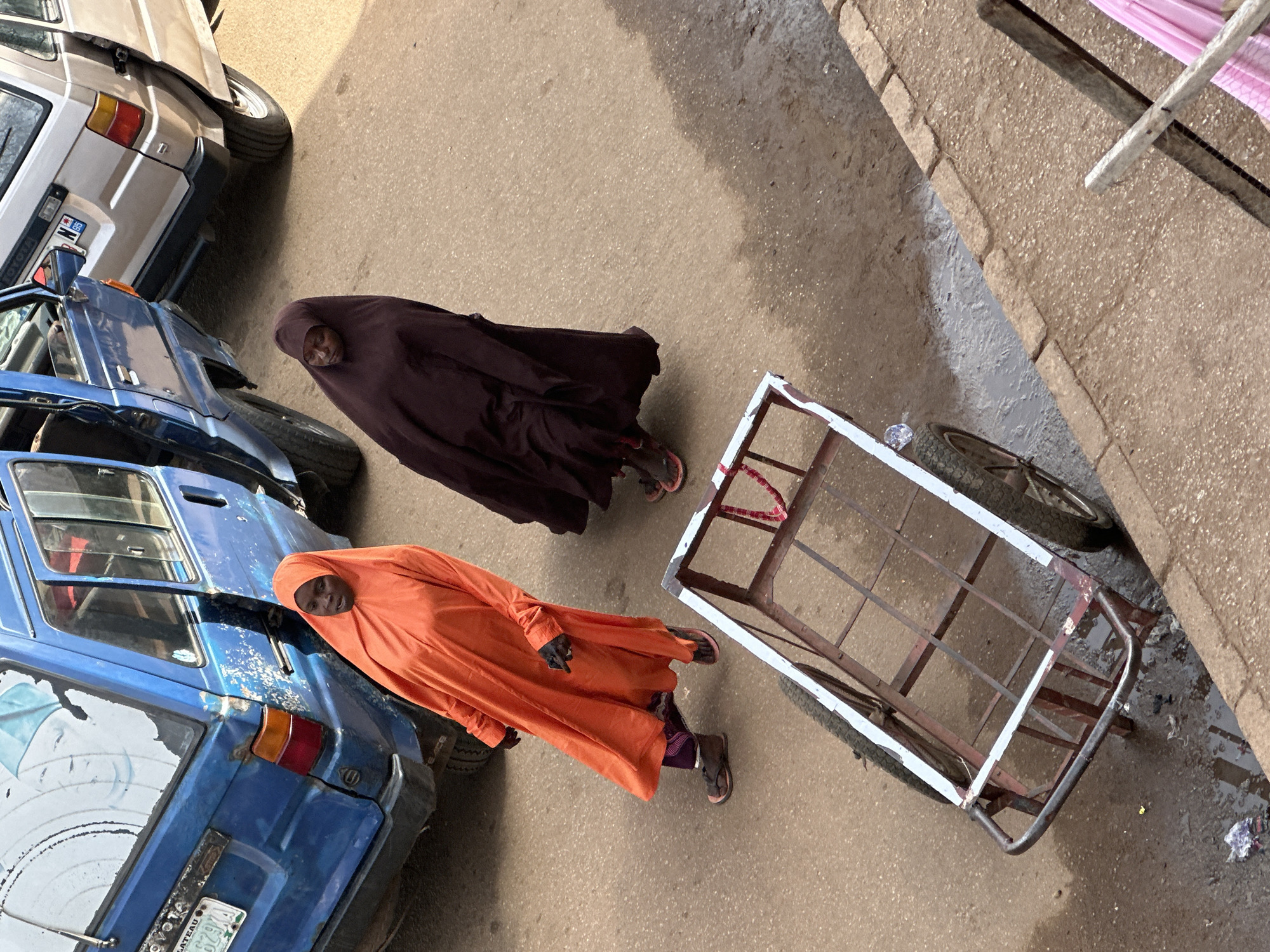 In this overhead shot, two individuals are standing on a paved surface, possibly a road or a courtyard, in what appears to be a marketplace or an area with parked vehicles. One person, dressed in a deep maroon hijab and covering, is positioned between two vehicles. The other individual, adorned in a vibrant orange hijab and covering, stands near a blue vehicle.
To the left of the frame, the rear of a light-colored van, possibly a Toyota, is visible. Beside it, a blue car, also showing signs of wear, is parked. In the foreground, a metal cart with a white frame and a cage-like structure is on the ground. The cart appears to be a hand-pulled trolley, possibly used for transporting goods.
The scene is illuminated by natural light, suggesting it is daytime. The presence of vehicles and the cart hints at a setting where trade or transportation might be common. The ground is a mix of asphalt and dirt, with some debris scattered around. The people's clothing, particularly the hijabs, suggests a cultural context where modest dress is observed. The overall impression is that of an ordinary day, possibly in a busy area of Bukuru, Nigeria. The image does not convey any specific emotions, but the subjects are captured in a static pose, as if paused during their activities. There is some visible text on a license plate of the blue vehicle.