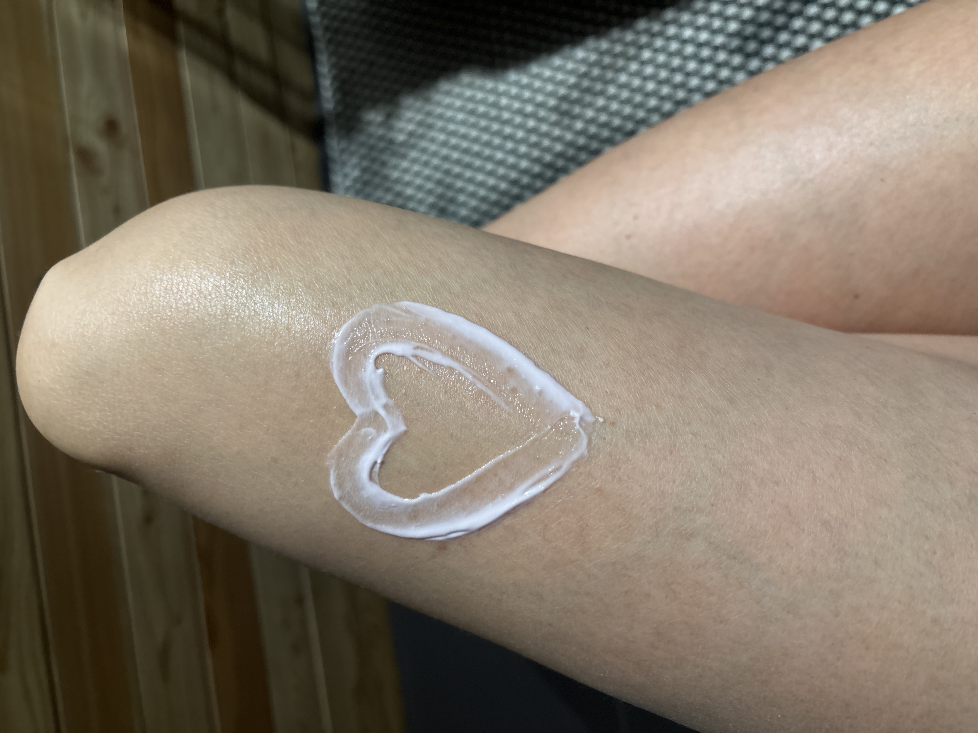 The image shows a close-up of a person's knee and lower thigh. A heart shape has been drawn on the skin using a white, creamy substance, possibly lotion or cream. The background is blurred, but it appears to consist of wooden panels on one side and a textured gray or black surface on the other. The location in Chaikovsky, Russia, is not apparent from the image.