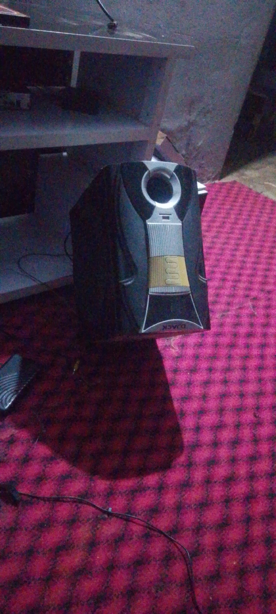 An indoor, low-angle, medium shot shows a black, upright speaker with a silver speaker grill and silver accent trim on a dark red, checkered carpet. A light grey shelf unit is visible in the background. A shadow of the speaker is cast on the carpet to the left. The scene is dimly lit, and the camera is out of focus. The speaker has the brand name "DJACK" printed on it in white lettering.
