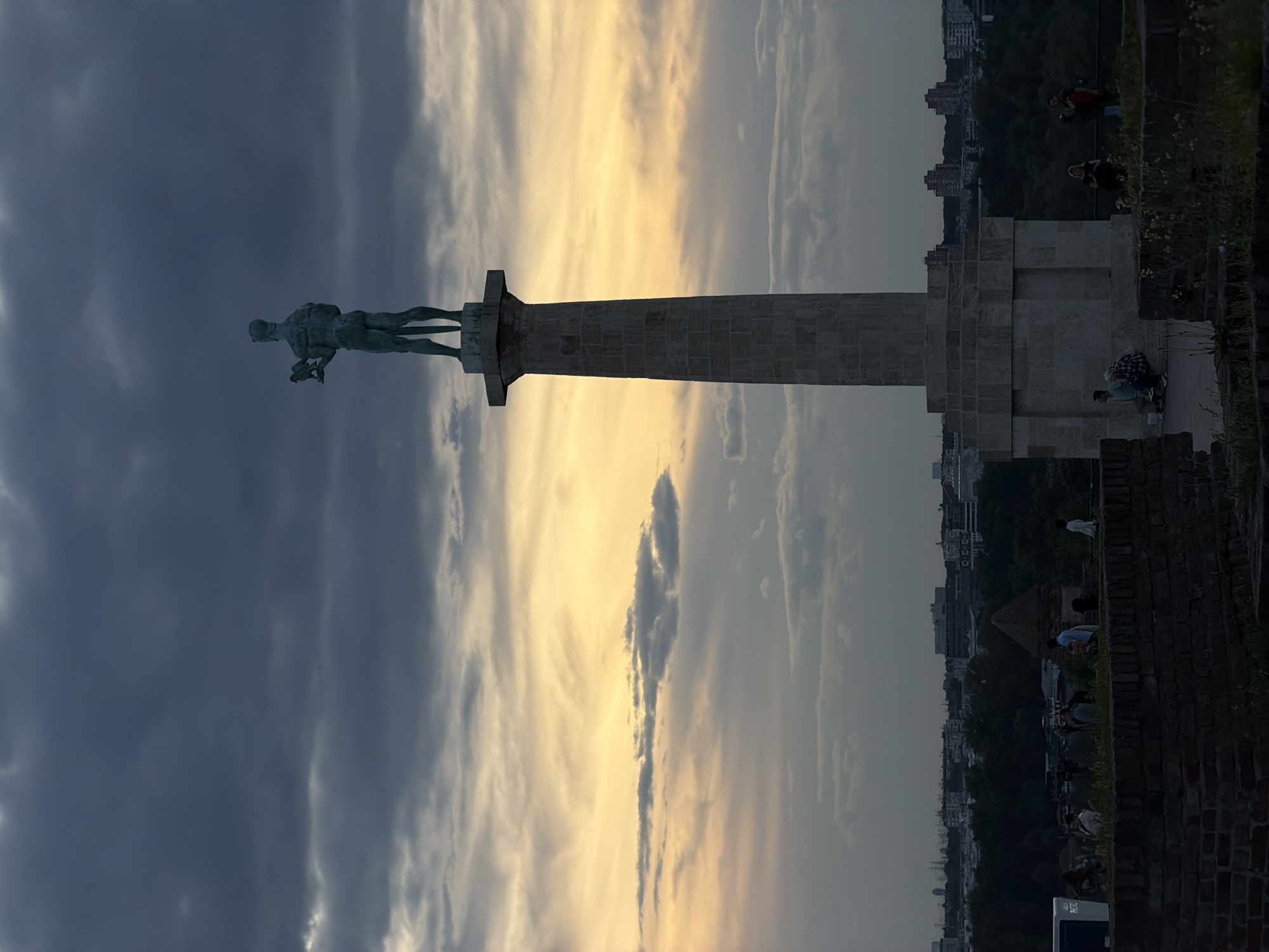 The image is of the Belgrade Victor monument in Belgrade, Serbia, silhouetted against a colorful sunset sky. The monument consists of a nude male figure holding a falcon, standing atop a tall column. The column has a square base made of light-colored stone blocks. A brick wall surrounds the base, and several people are present on the wall, some standing and some sitting. Beyond the wall, there are buildings and trees. The sky transitions from dark, stormy clouds to yellow and orange hues near the horizon.