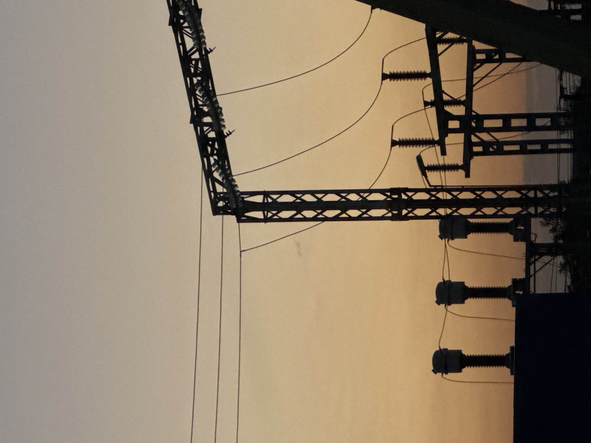 The image shows a silhouette of an electrical substation against a gradient sky of pale orange and beige, suggesting either sunrise or sunset. Three large, ribbed insulators are prominently visible in the foreground, mounted on a dark, low structure. Behind these, a tall, lattice-style metal tower supports a horizontal beam with multiple smaller insulators. Several power lines are strung between these structures. A diagonal beam of a larger structure enters from the right side of the frame, obscuring some of the background. The overall scene is dark and industrial, with no visible people or specific urban landmarks.