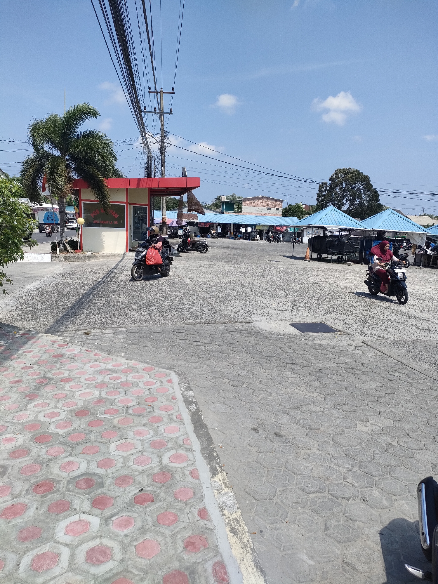 The image depicts an outdoor scene in the city of Sungailiat, Indonesia, likely a marketplace or a transport hub, with a bright, sunny sky. A red and white building, possibly a small office or shop, is visible on the left, with a palm tree in front of it. Various market stalls with blue awnings are set up in the background. Several people are present, predominantly riding motorcycles. One individual in a red headscarf is riding a black motorcycle in the foreground on the right. Another person on a black motorcycle with a red bag is in the middle ground on the left. The ground is paved with hexagonal interlocking concrete blocks, with a section of similar blocks featuring pink accents in the lower left corner. Electrical wires are prominently strung across the sky.