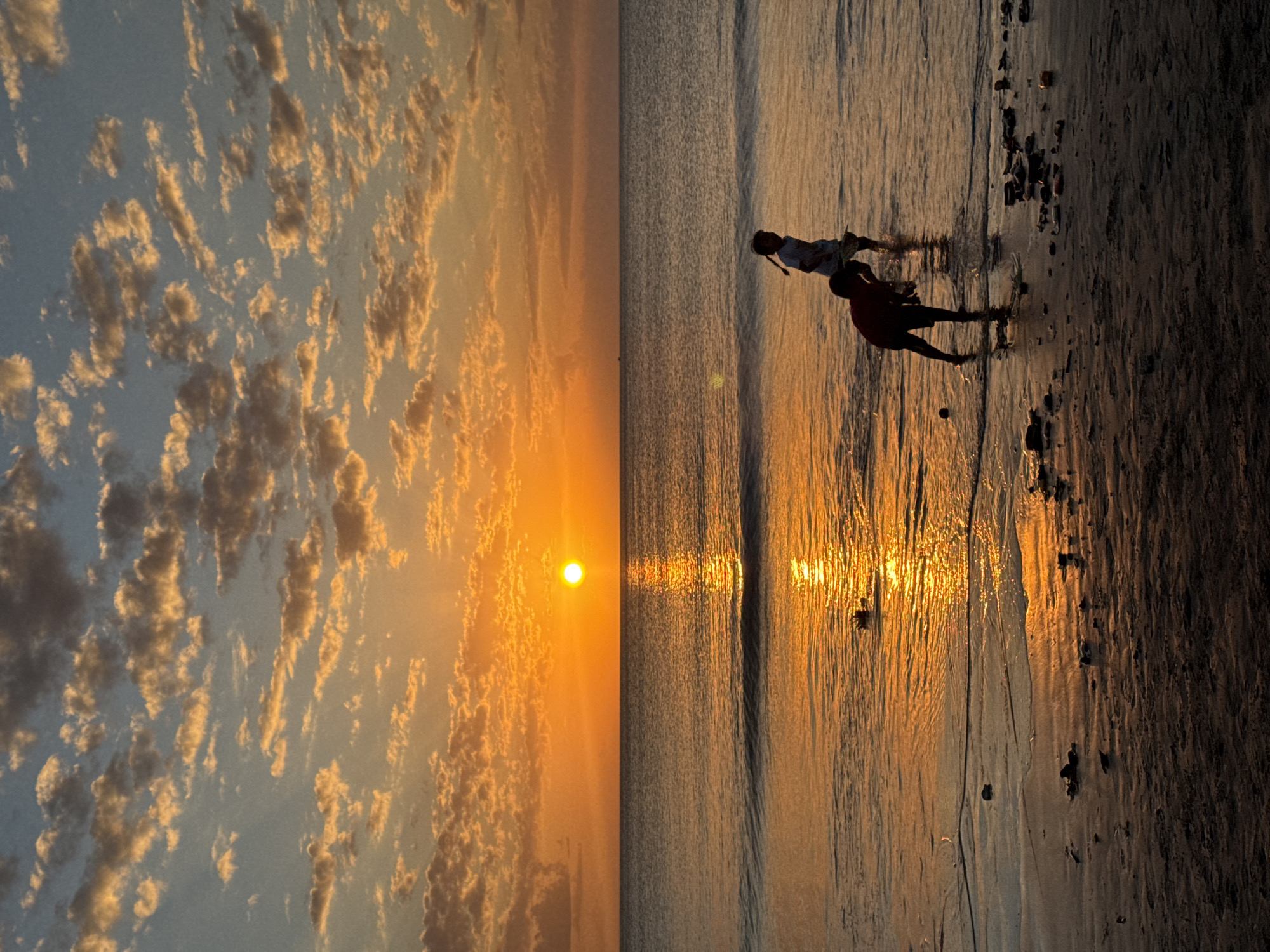 A sunset on the beach in Kuta Selatan, Indonesia, casts a warm glow over the ocean and the sky. The sun, a bright orange orb, is partially obscured by scattered clouds tinged with orange and yellow. The ocean is calm with gentle waves lapping at the shore. Two children are silhouetted against the vibrant sunset as they play in the shallow water. The beach is wet sand, littered with debris, and reflects the warm hues of the sky.