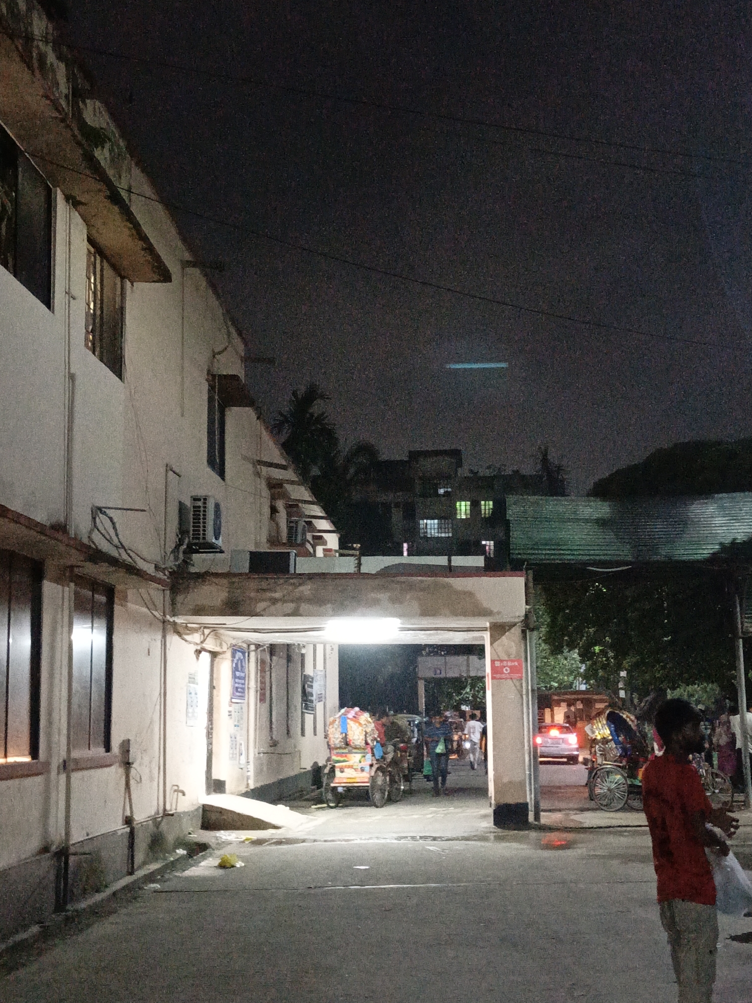 The image shows a nighttime street scene in an urban area of Unknown, Unknown. A light-colored building is on the left side of the frame, and it has windows and an air conditioning unit. The street is paved and leads to a covered area with lights. Several people are present in the covered area, some walking, and others are near rickshaws. A man wearing a red shirt and light-colored pants stands on the right side of the frame, partially obscuring the view of the street beyond. There are vehicles and trees visible in the background. The sky is dark, suggesting it is nighttime.