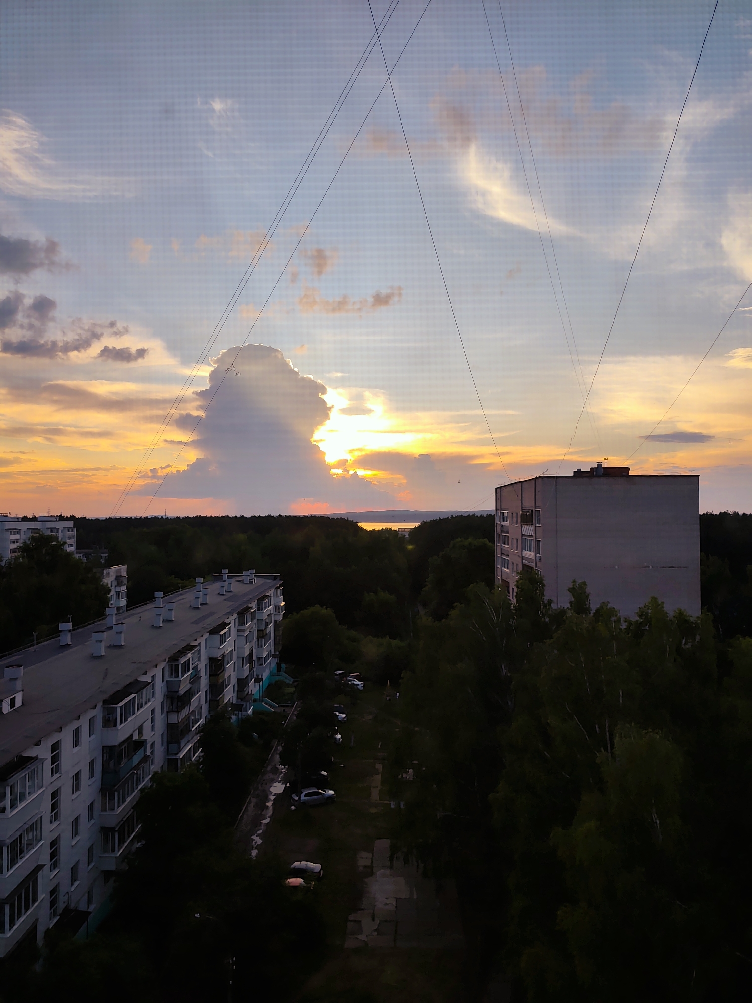 This is a high-angle, slightly downward-facing medium shot of a residential area in Chaikovsky, Russia, during a sunset. The sky is a vibrant mix of orange, yellow, and blue, with scattered clouds. A large, prominent cloud formation is illuminated by the setting sun, casting rays of light. Several power lines crisscross the sky diagonally.
In the foreground, on the left, is a long, multi-story apartment building with a grey roof. The building has numerous balconies with railings, and white walls. To the right of this building, in the midground, is a densely wooded area that extends to the horizon. Further into the midground and to the right, another, taller, plain grey apartment building stands. The ground level is obscured by trees and the shadows of the buildings. A street or path runs between the buildings and the wooded area, with parked cars visible, some appearing wet or reflecting light. The overall environment appears to be a typical Soviet-era residential district surrounded by green spaces. No people are visible in the frame.