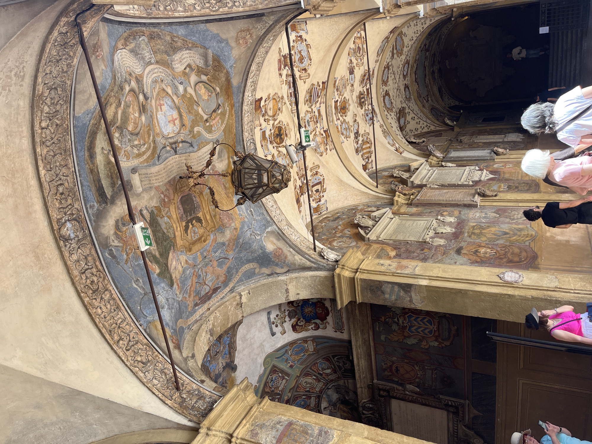 The image depicts the ornate, mural-covered passageway of Palazzo d'Accursio in Bologna, Italy. The vaulted ceiling features numerous painted coats of arms and decorative elements. A hanging lantern is prominently displayed. Several people are visible, some seemingly viewing the murals and architectural details. The scene suggests a historical and artistic setting, typical of the Palazzo d'Accursio's historical significance.