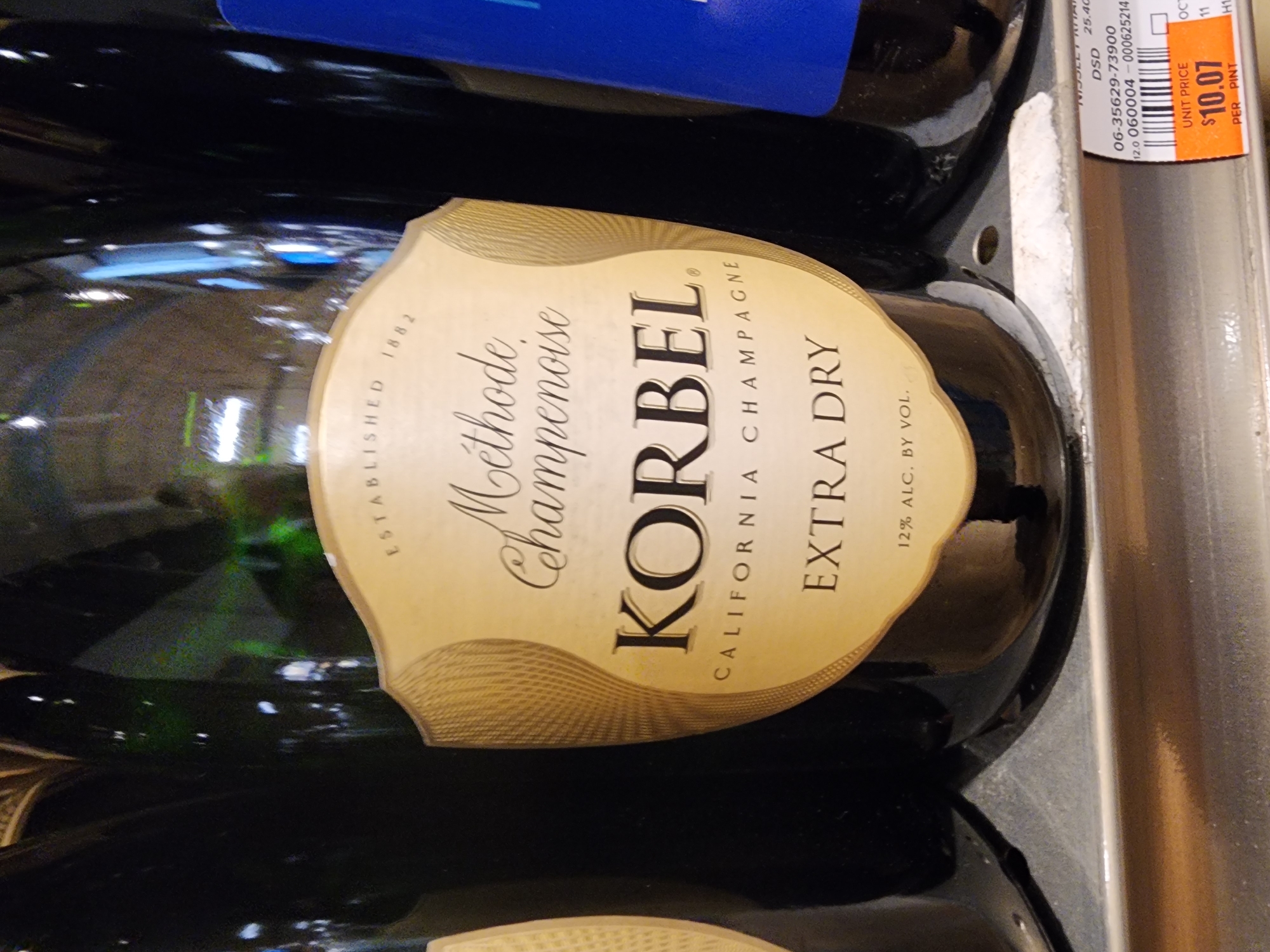 A close-up shot captures a bottle of Korbel California Champagne, Extra Dry, positioned on a shelf. The bottle has a gold label with embossed text that reads "ESTABLISHED 1882," "Méthode Champenoise," "KORBEL," "CALIFORNIA CHAMPAGNE," and "EXTRA DRY." Below "EXTRA DRY," it states "12% ALC. BY VOL." The bottle is dark glass. To the right of the Korbel bottle, another bottle with a blue label is partially visible. The background is dark, suggesting indoor lighting. In the lower right corner, a price tag with "UNIT PRICE $10.07 PER PINT" is visible. The scene appears to be in a retail environment, likely a store.