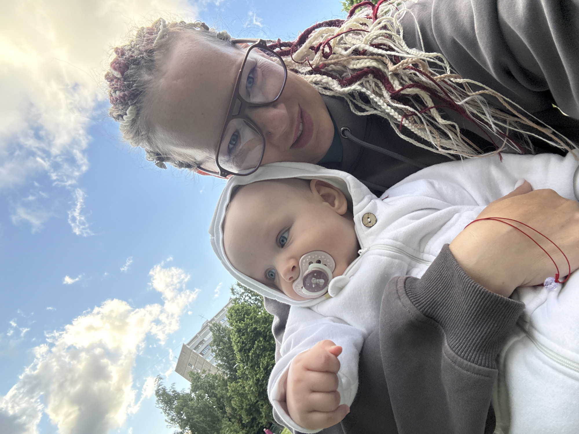 A selfie taken outdoors on a sunny day with a blue sky and scattered clouds. The photographer, a woman with blonde and burgundy braids, wears glasses and a dark hoodie. She is holding a baby, who is wearing a white hooded outfit with a pacifier in its mouth. In the background, there are trees and a multi-story apartment building, suggesting an urban residential area. The lighting and clouds indicate it is likely daytime.