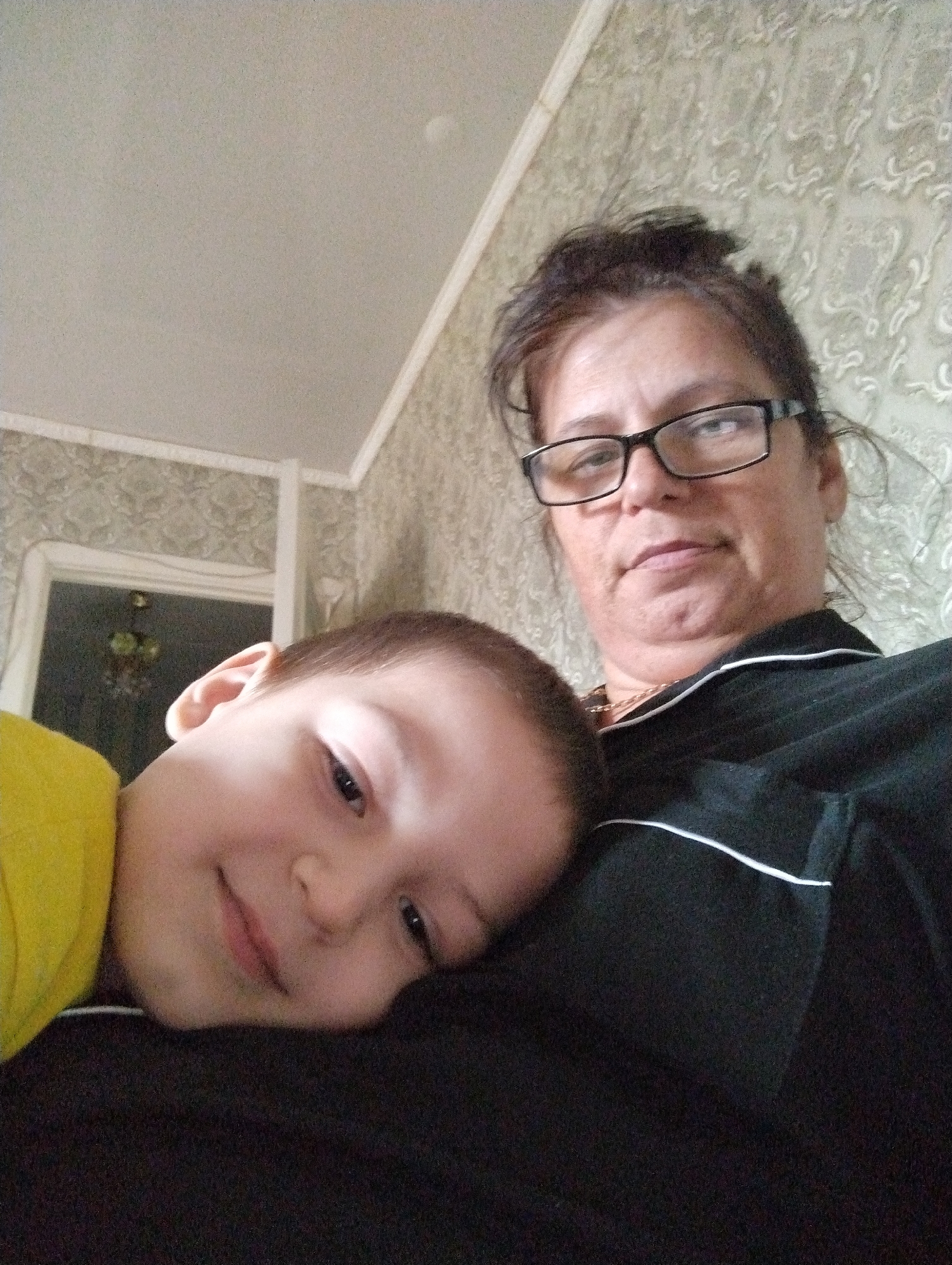 A woman and a child are taking a selfie in an indoor setting. The woman is wearing glasses and a black shirt with white trim. The child, wearing a yellow shirt, is resting their head on the woman's shoulder. The background appears to be a room with patterned wallpaper and a ceiling. The image is taken from a low angle, looking upwards. The location is Kingisepp, Russia.