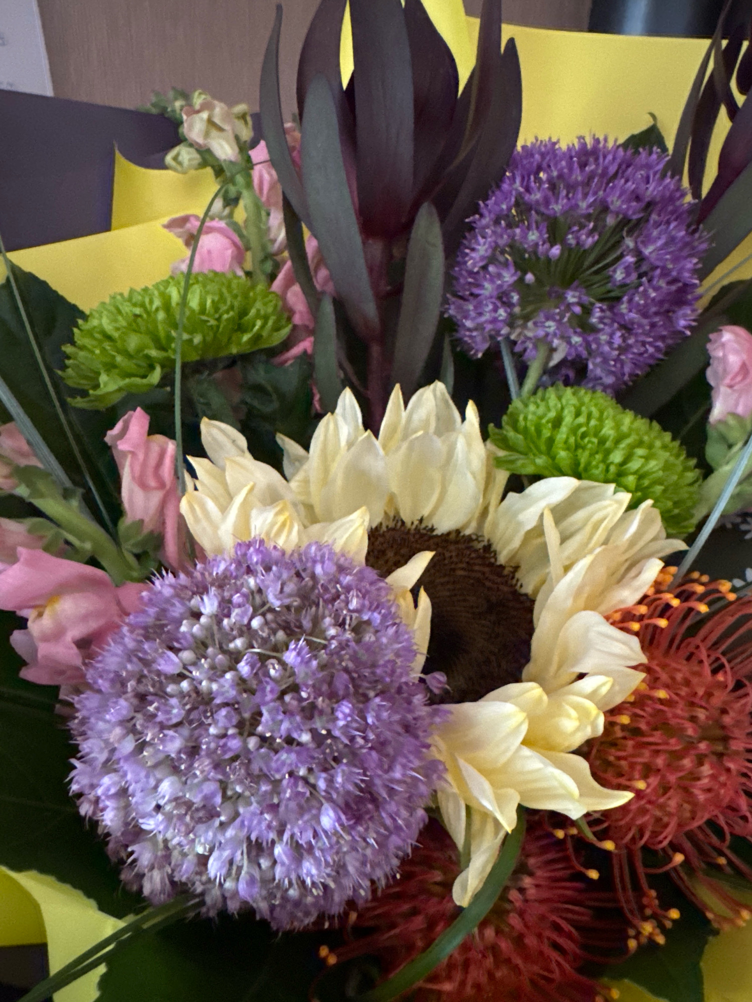 The image shows a close-up of a bouquet of flowers. The bouquet contains a variety of colorful blooms including a large sunflower with pale yellow petals and a dark brown center, two spherical purple allium flowers, a cluster of small green chrysanthemums, and some red pincushion proteas. There are also some pink snapdragon flowers and dark purple leaves or petals from a protea. The flowers are arranged against a backdrop of yellow and dark purple wrapping paper. The scene is indoors, with the lighting suggesting it is natural light. There are no people or animals visible in the image. The image does not contain any information that would allow for the determination of its location in the city of Захарьино, Russia.