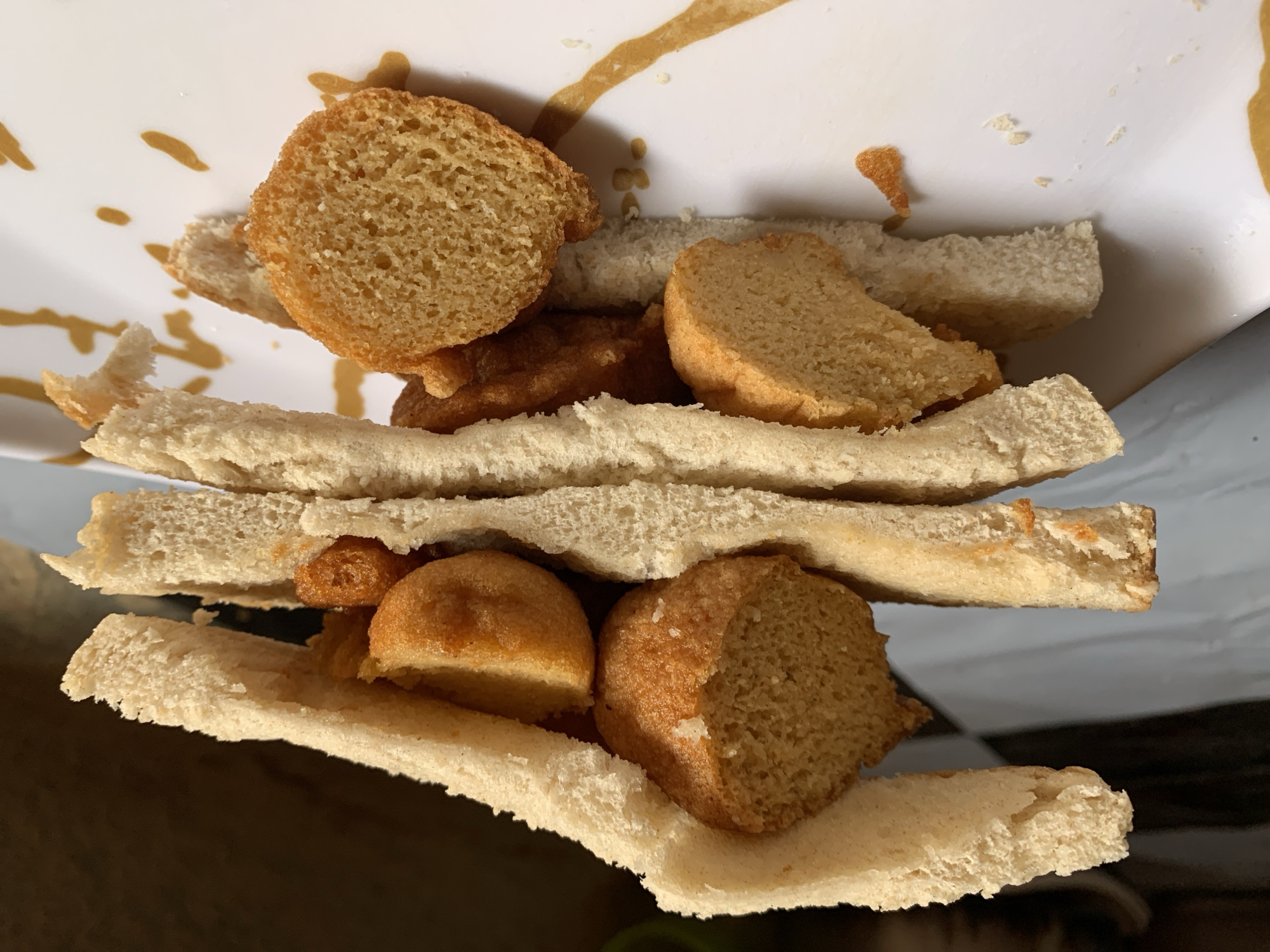 A close-up, slightly tilted shot depicts a homemade sandwich or similar food item. The sandwich is assembled with slices of white bread, with a variety of round, toasted bread pieces between the slices. Some of the toasted bread pieces are golden brown, while others are lighter. There is a streak of brownish liquid on the white plate the sandwich is on. The plate appears to be resting on a darker surface. The location is not discernible from the image.