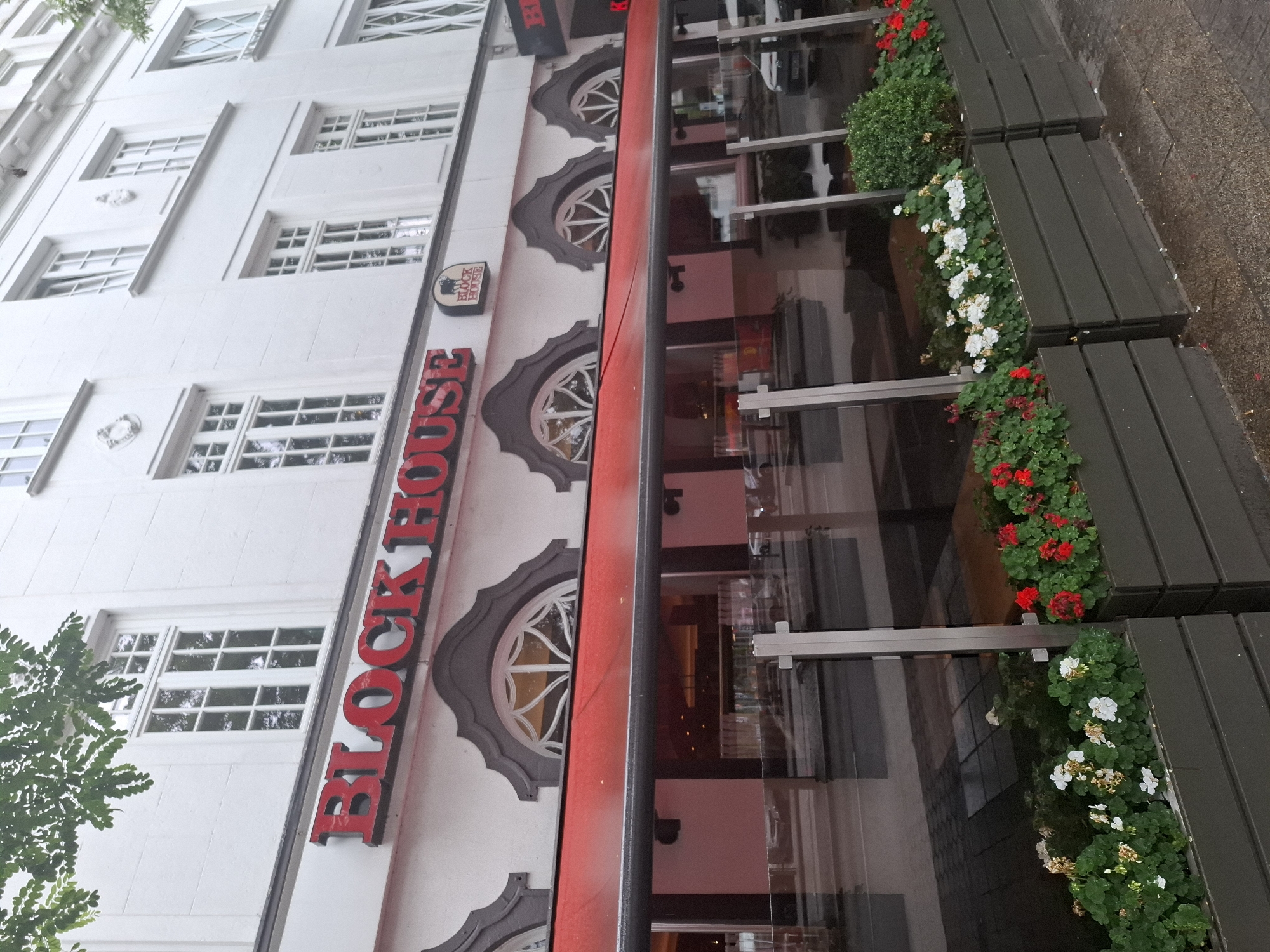 The image shows the facade of a restaurant called "BLOCK HOUSE" in Hamburg, Germany. The restaurant has a red awning over its windows and outdoor seating area. There are planters with red and white flowers in front of the restaurant. The building is white with several windows on the upper floors. The street in front of the restaurant is wet, indicating it may have recently rained. There are no people visible in the image.